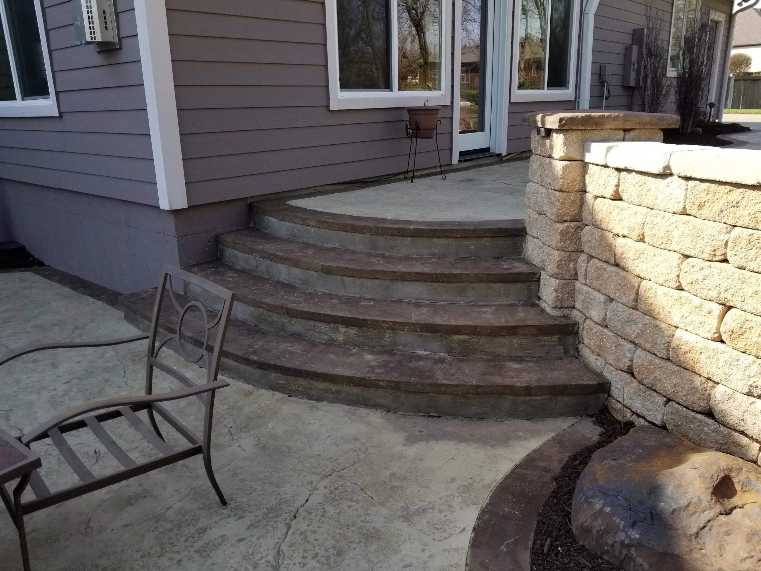 Exterior view of a house with a curved stone staircase leading to a sliding glass door. To the right, there is a beige stone retaining wall with a large rock at its base. A metal chair with armrests is on the concrete patio in the foreground. The hou