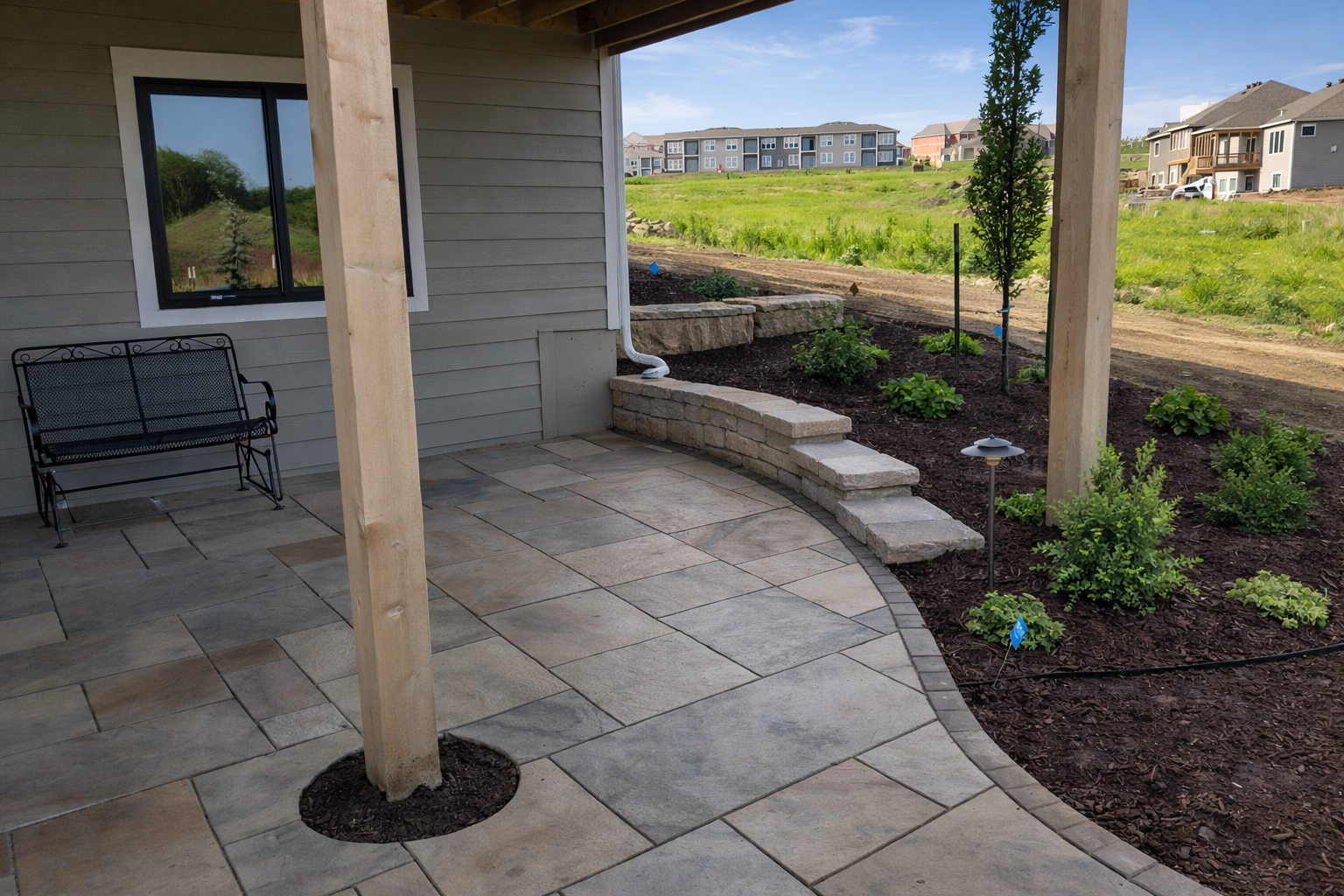 Back porch area with stone pavers, a black metal bench, a wooden support post, a garden bed with small shrubs, a young tree, and landscape lighting, overlooking a grassy field and neighboring houses.