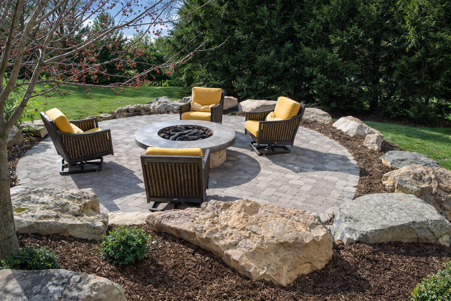 Outdoor patio with four black wicker chairs with yellow cushions arranged around a circular stone fire pit on a circular paved area surrounded by rocks and landscaping plants.