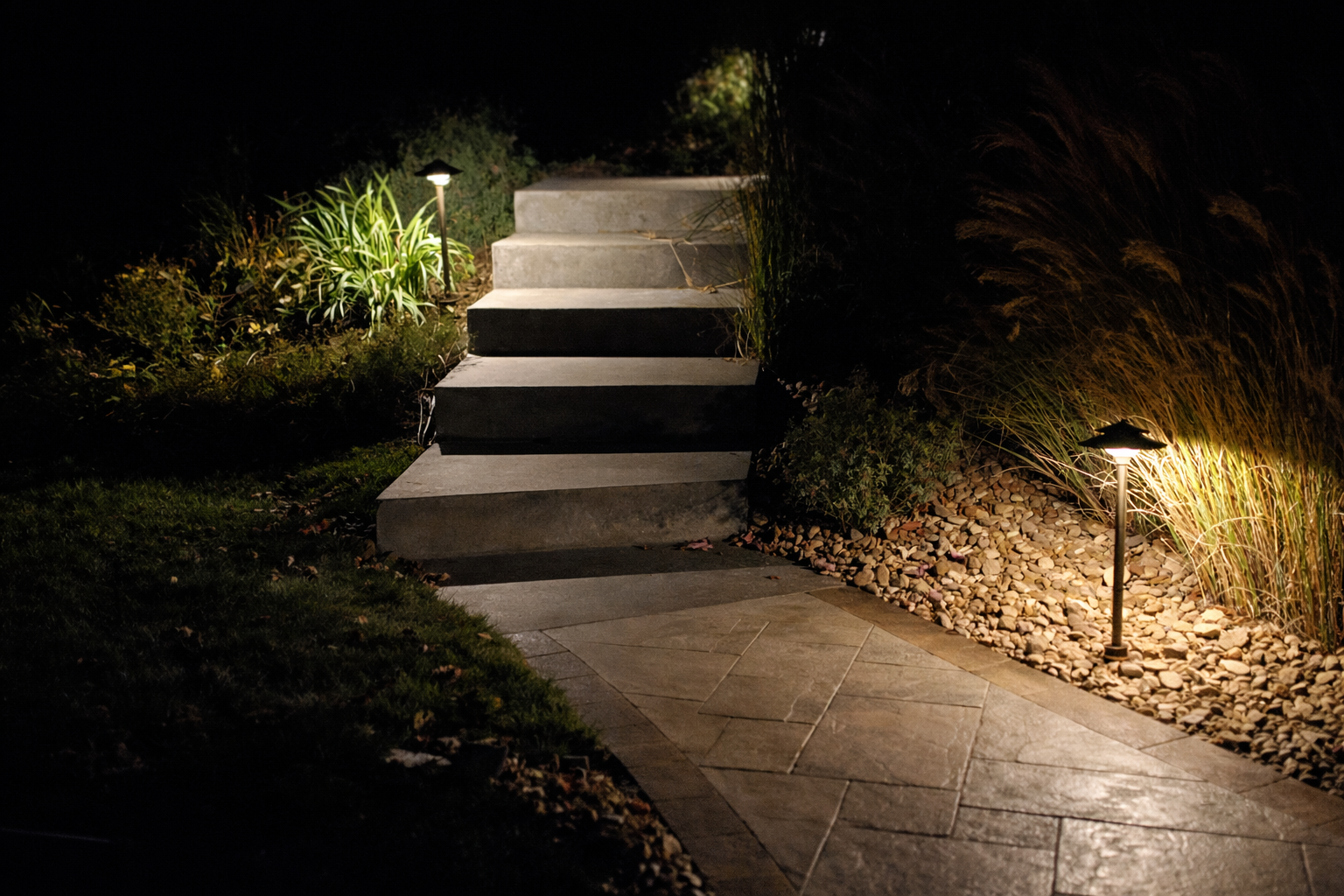 Nighttime outdoor scene featuring a concrete staircase with five steps, illuminated by ground-level pathway lights on either side, with landscaped plants and gravel surrounding the stairs and walkway.