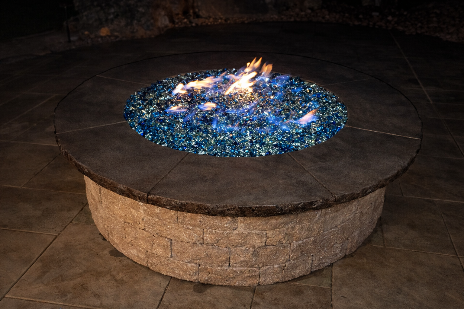 A stone fire pit with a dark top and a circular arrangement of blue decorative glass stones inside, with flames rising from the glass.