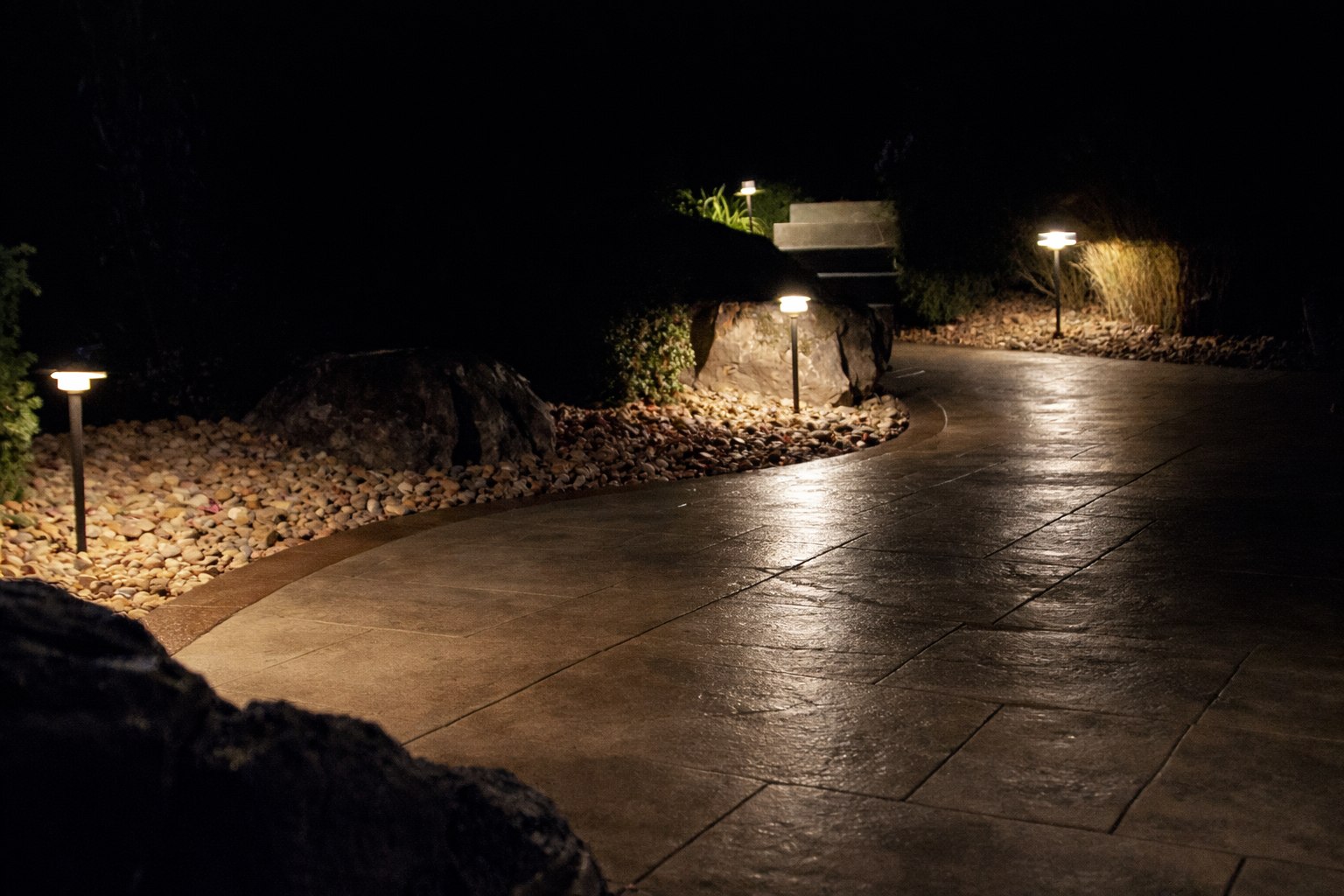 A winding outdoor pathway illuminated by small landscape lights at night, with rocky borders and lush greenery.