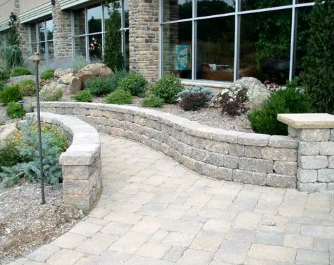 Curved stone retaining wall with landscaping and pathway in front of a building with large windows.
