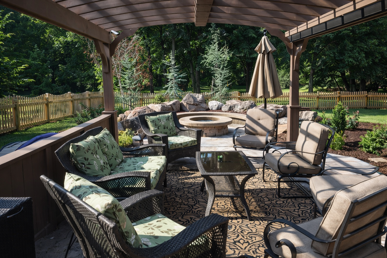 A backyard patio with outdoor furniture, including cushioned chairs, a glass-top table, and a large umbrella, under a wooden pergola. There is a fire pit with rocks and trees in the background.