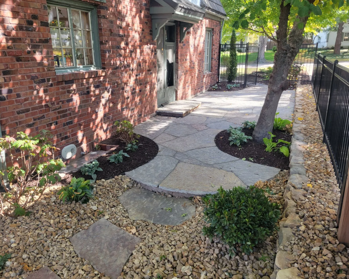 A small backyard garden with a brick house, stone pathway, trees, and plants surrounded by gravel and a black fence.