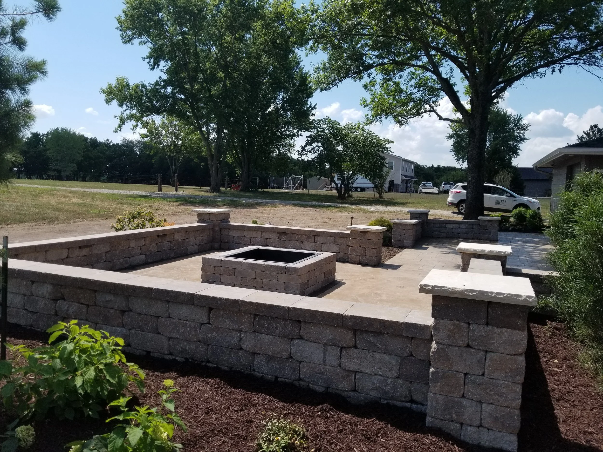 A stone fire pit surrounded by a low stone wall and benches in a backyard with trees, a house, and parked cars in the background.