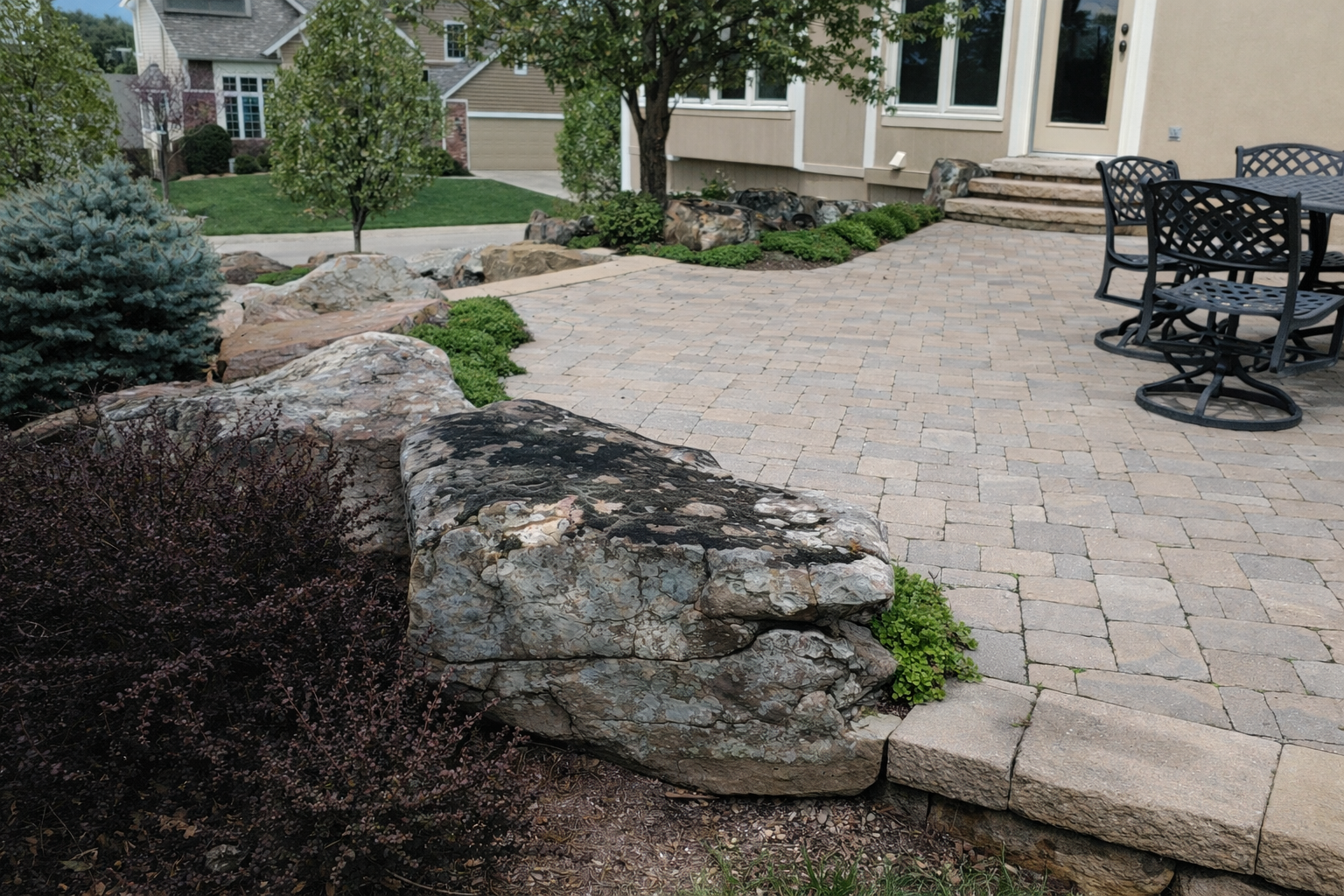 A landscaped backyard with a stone patio, large rocks, small trees, shrubs, and outdoor furniture.