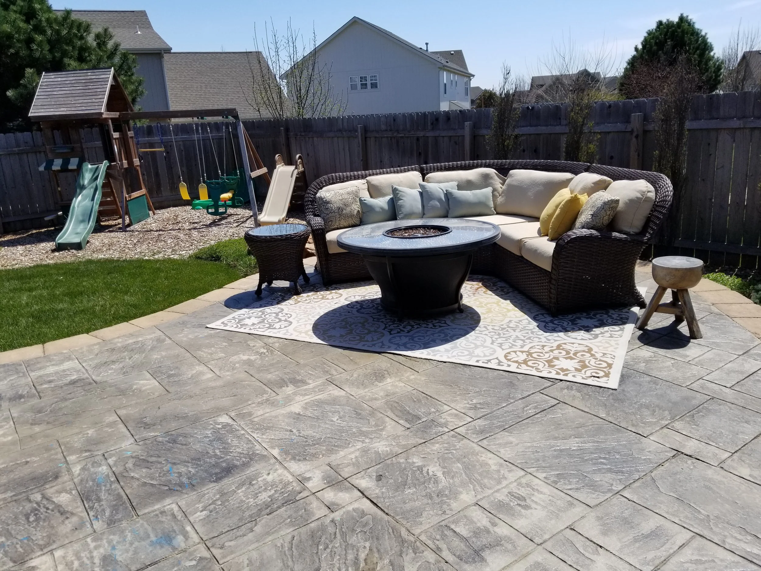 Backyard patio with a large wicker sectional sofa with beige cushions and throw pillows, a round fire pit table, a small stool, and a play area with a swing set, slide, and sandbox in the background, enclosed by a wooden fence.