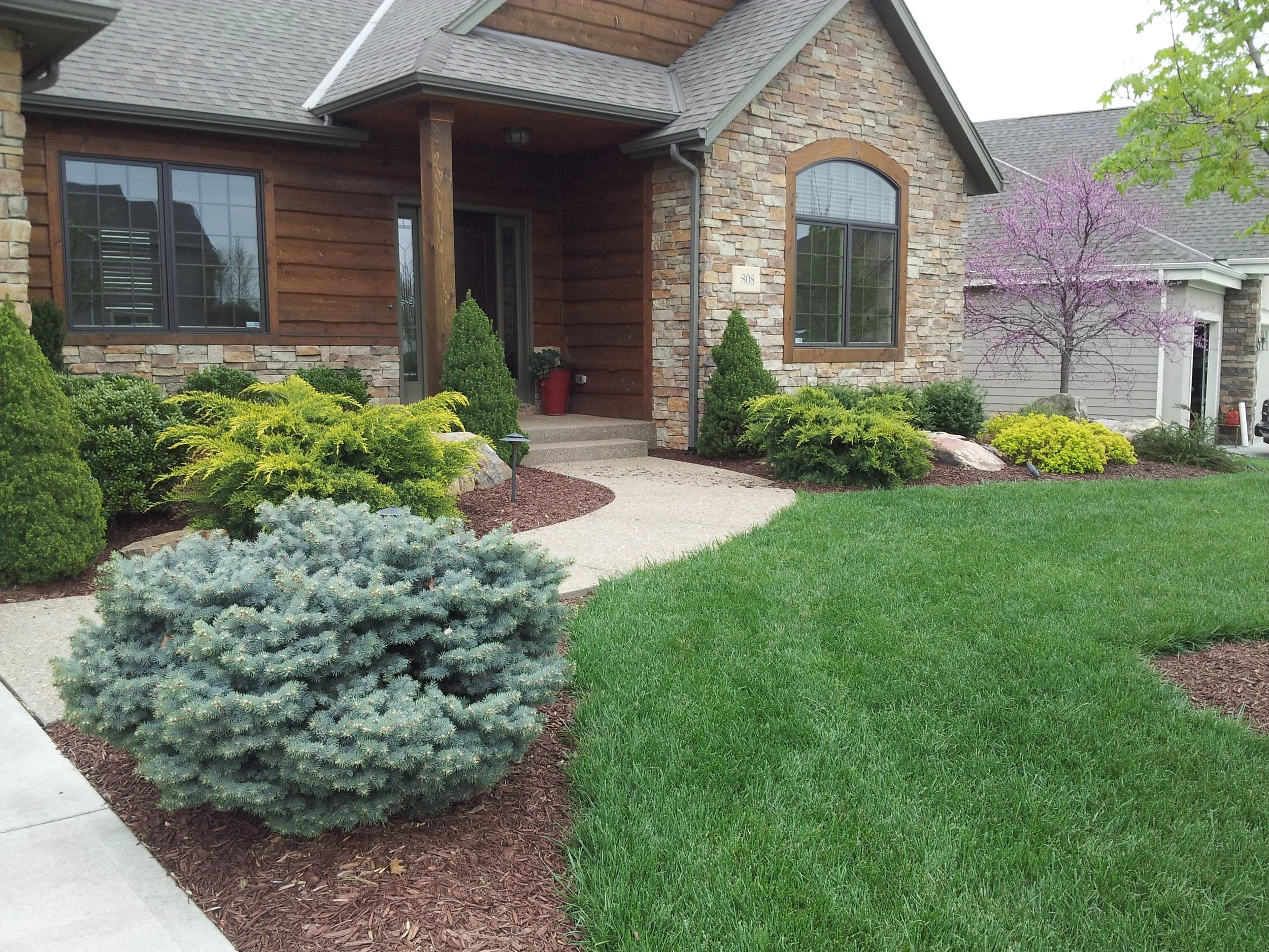 Front yard with green grass, shrubs, and a house with a stone and wood exterior, including a porch and a purple flowering tree in the background.