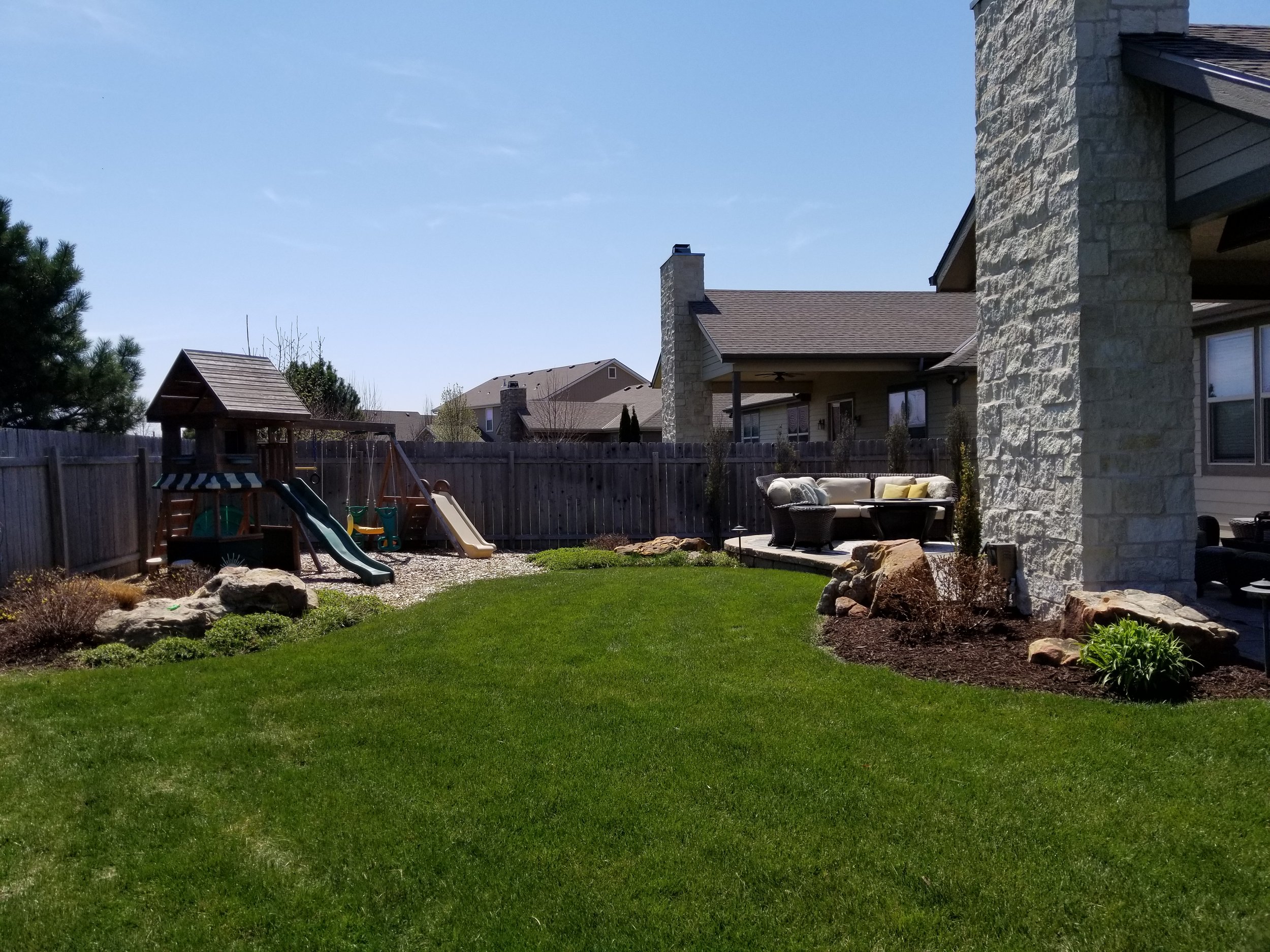 Backyard with a wooden playset and slide, outdoor seating area, green lawn, landscaped garden beds with rocks and plants, wooden fence, neighboring houses, and a blue sky.