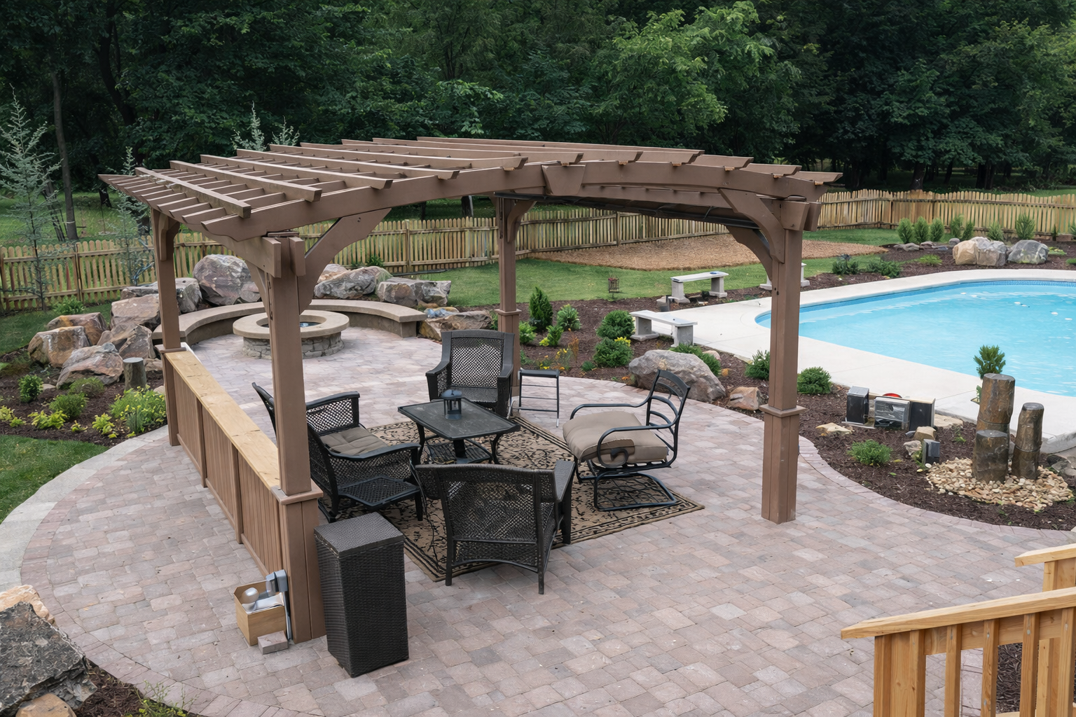 Backyard patio with a brown pergola, black outdoor furniture, a fire pit, landscaping with rocks and bushes, a swimming pool, and a wooden fence