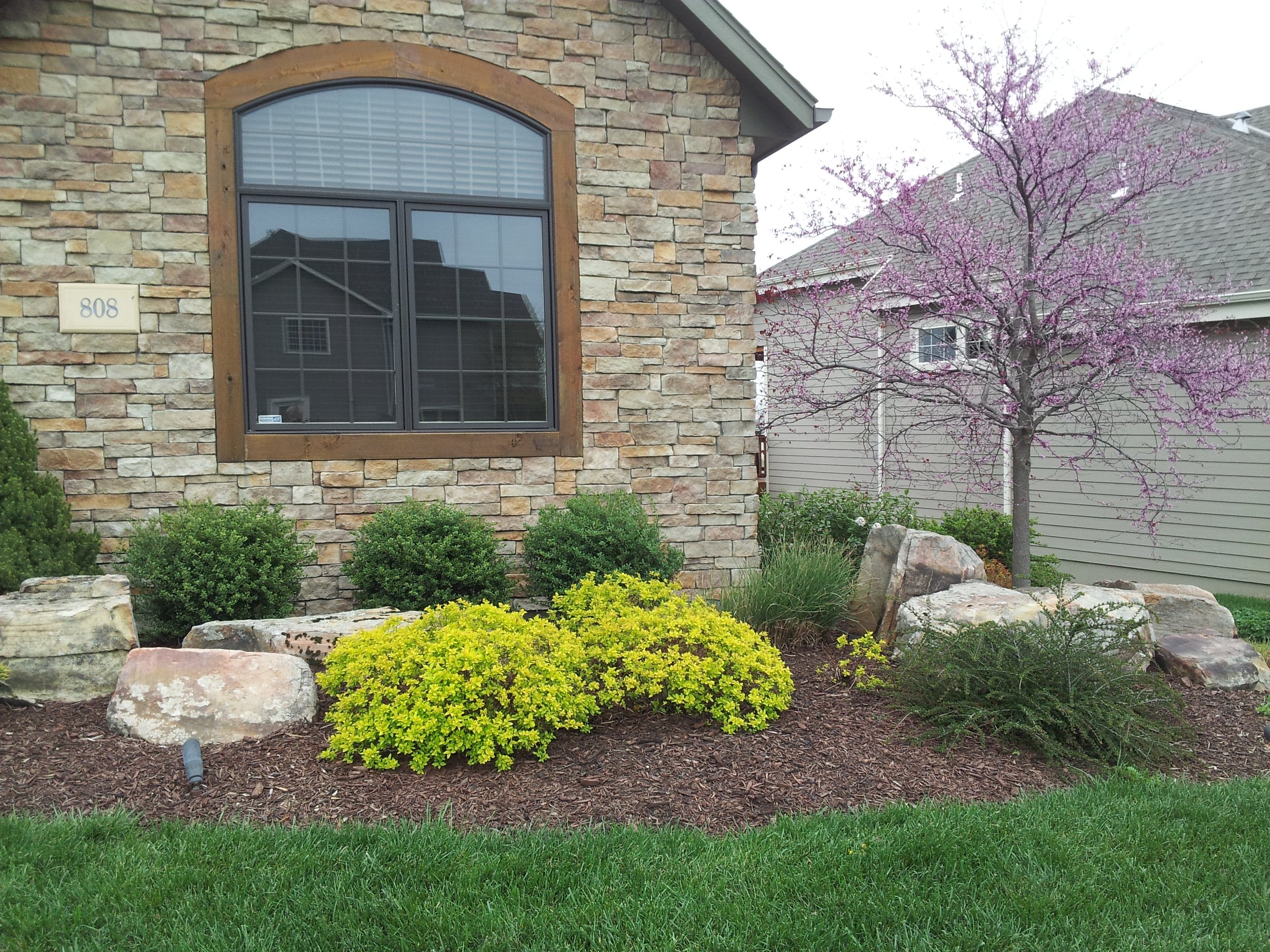 View of a house with a stone facade, a large arched window with wooden framing, and a landscaped front yard with bushes, large rocks, and a flowering tree with pink blossoms.