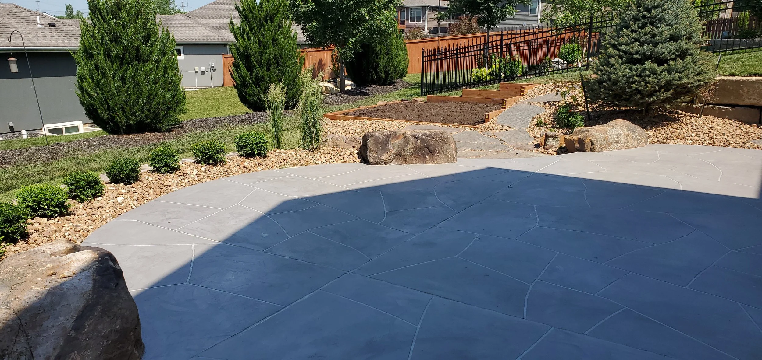 A backyard with a stamped concrete patio, small bushes in mulch beds, large rocks, and a winding stone pathway leading to a small deck and garden area with a black metal fence, evergreen trees, and a grassy lawn.