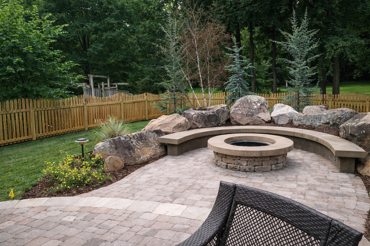 Backyard patio with a fire pit, curved concrete bench, large rocks, and surrounding trees and plants, enclosed with a wooden fence.