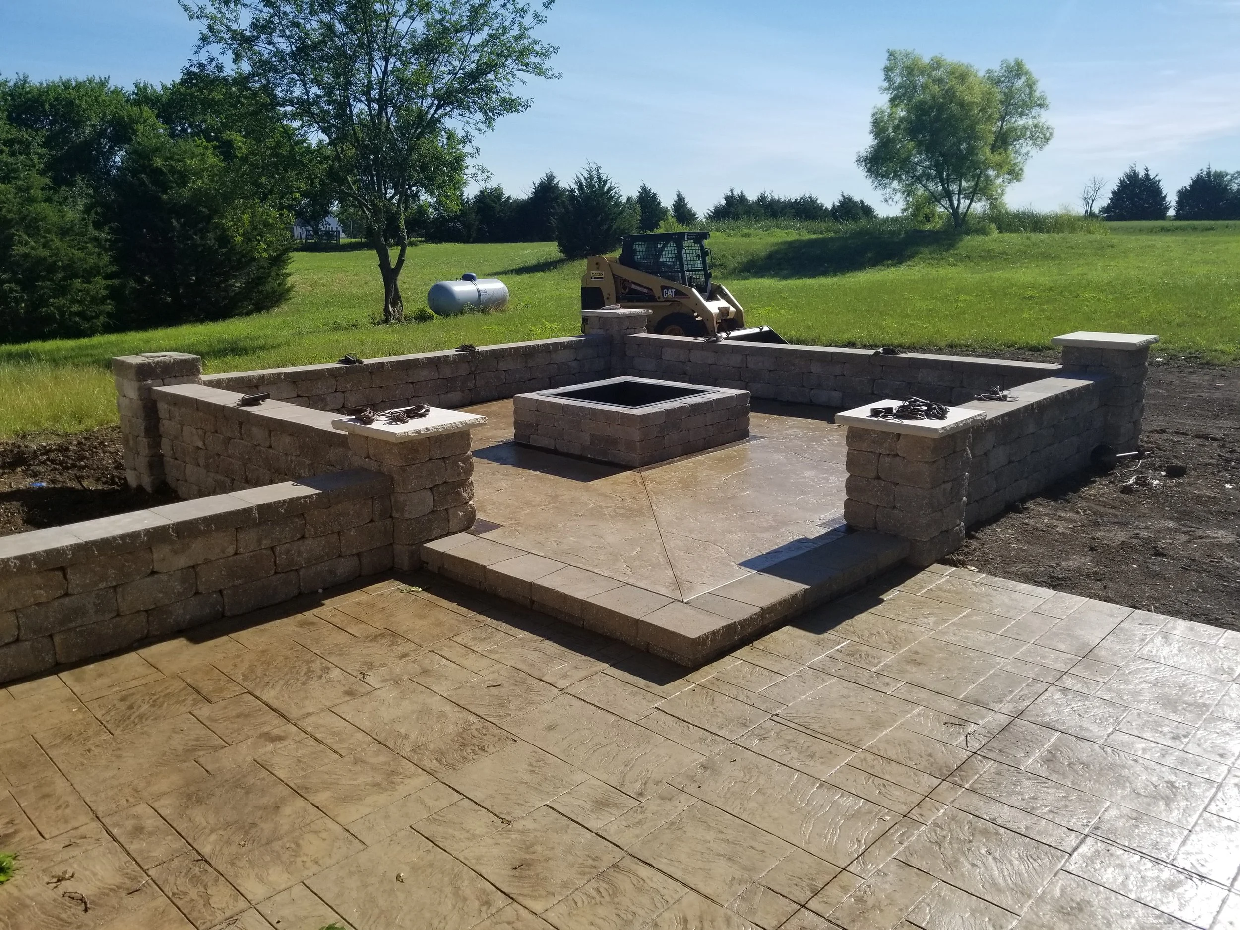 A newly constructed outdoor stone patio and fire pit area with low stone walls, surrounded by grass and trees, and construction equipment in the background.