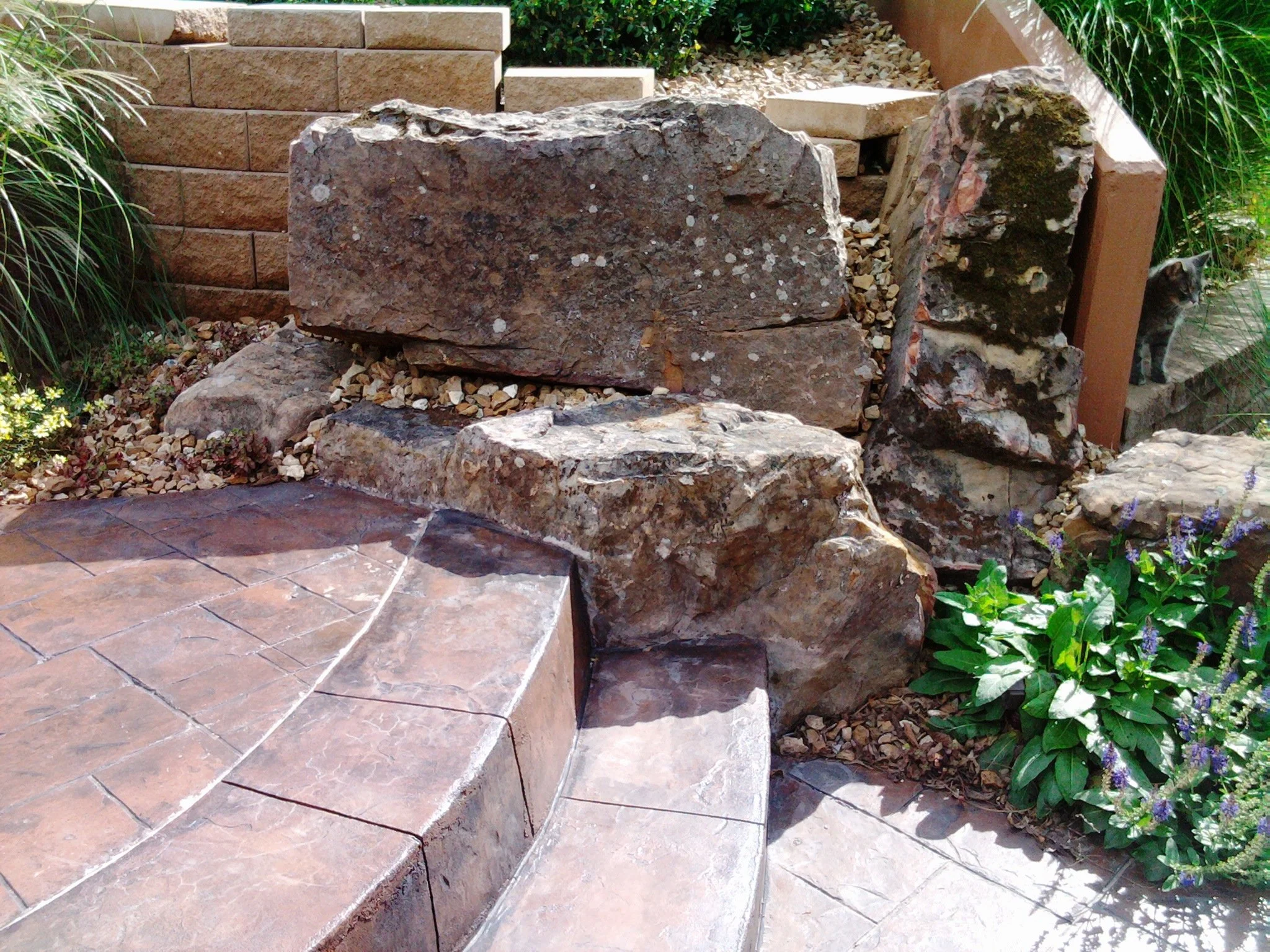 Landscape with large rocks, brick pathway, and green plants with purple flowers.