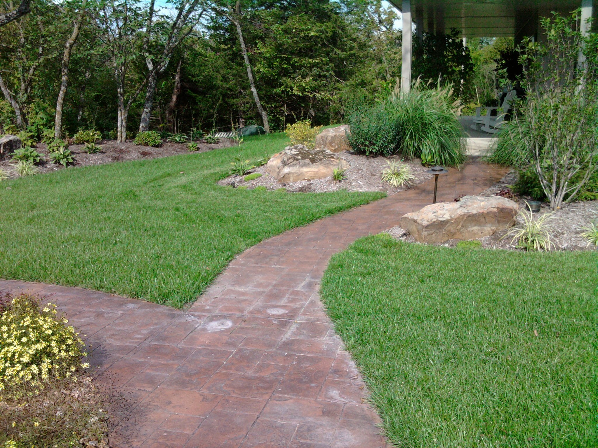 Curved brick pathway running through a well-maintained lawn and garden with trees, shrubs, and rocks.
