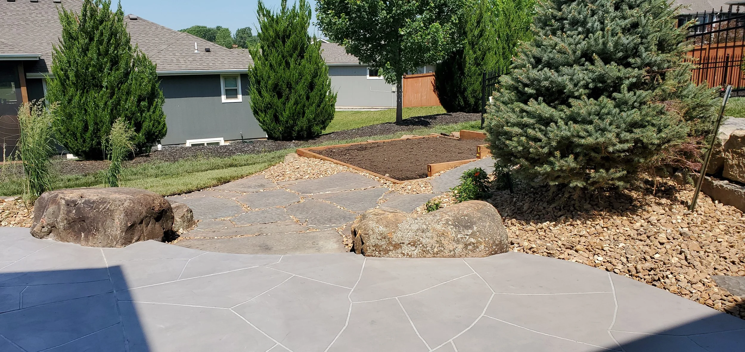 Backyard garden with large rocks, gravel, mulch, and freshly turned soil in a wooden frame bed. Surrounding greenery includes shrubs, trees, and grass. Part of the patio or walkway is visible in the foreground with a shadow cast over it.