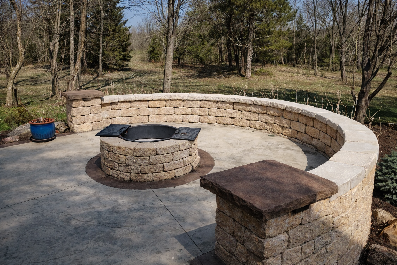 Outdoor patio with circular stone fire pit and curved stone wall, surrounded by trees and grass.