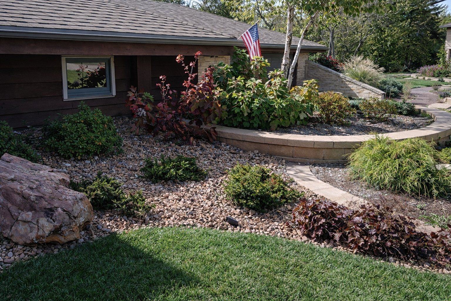 A landscaped backyard with a variety of plants, a curved stone retaining wall, a flag on a pole, and a window on a brown house. The yard features gravel, grass, and a stone pathway, with trees and shrubs in the background.