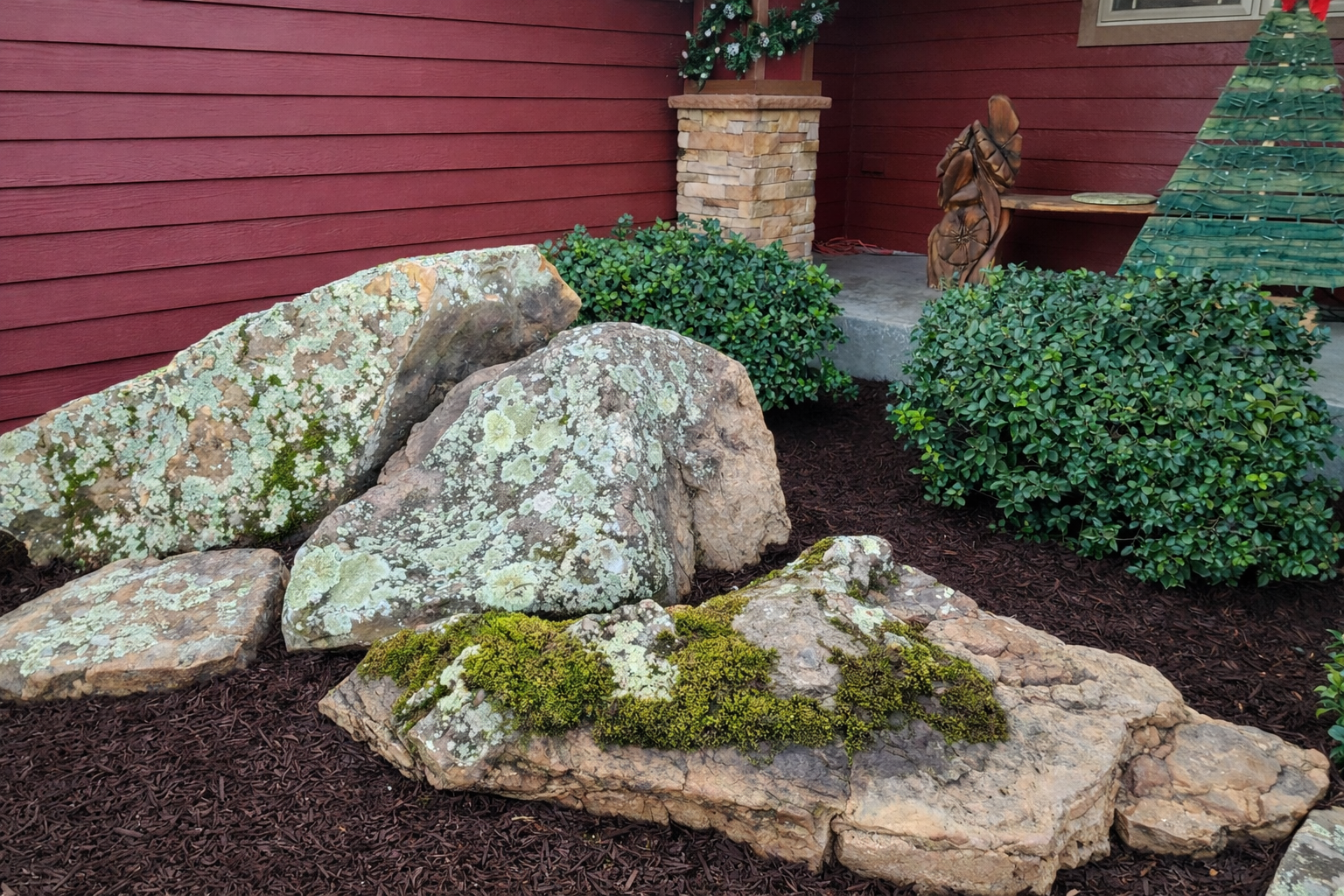 Decorative garden with large rocks covered in lichen and moss, green bushes, mulch ground, red wooden wall, and a small statue of an angel near a wooden table decorated with Christmas ornaments.
