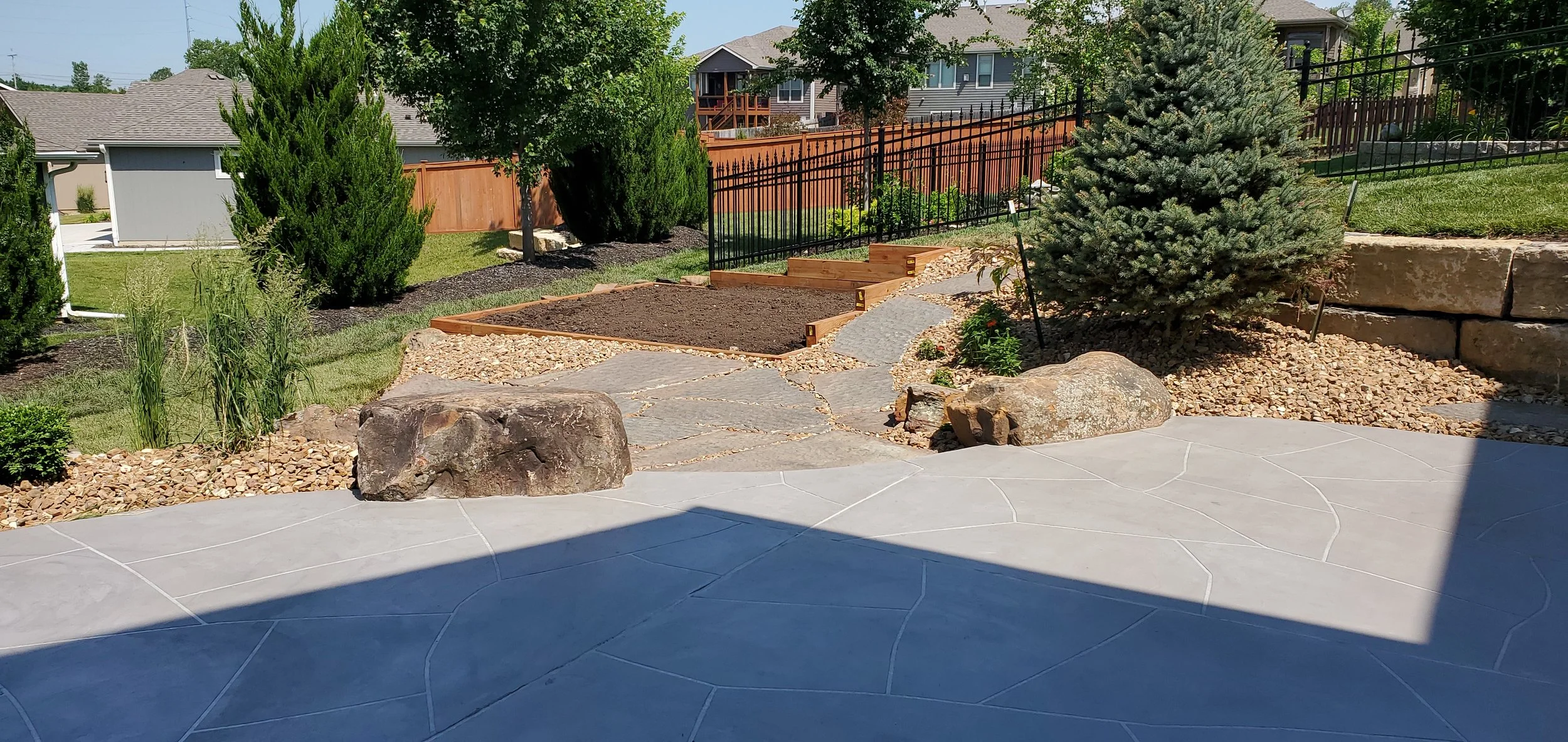 Backyard garden with stone patio, large rocks, trees, shrubs, and a fenced yard with stairs leading to a higher level of ground.