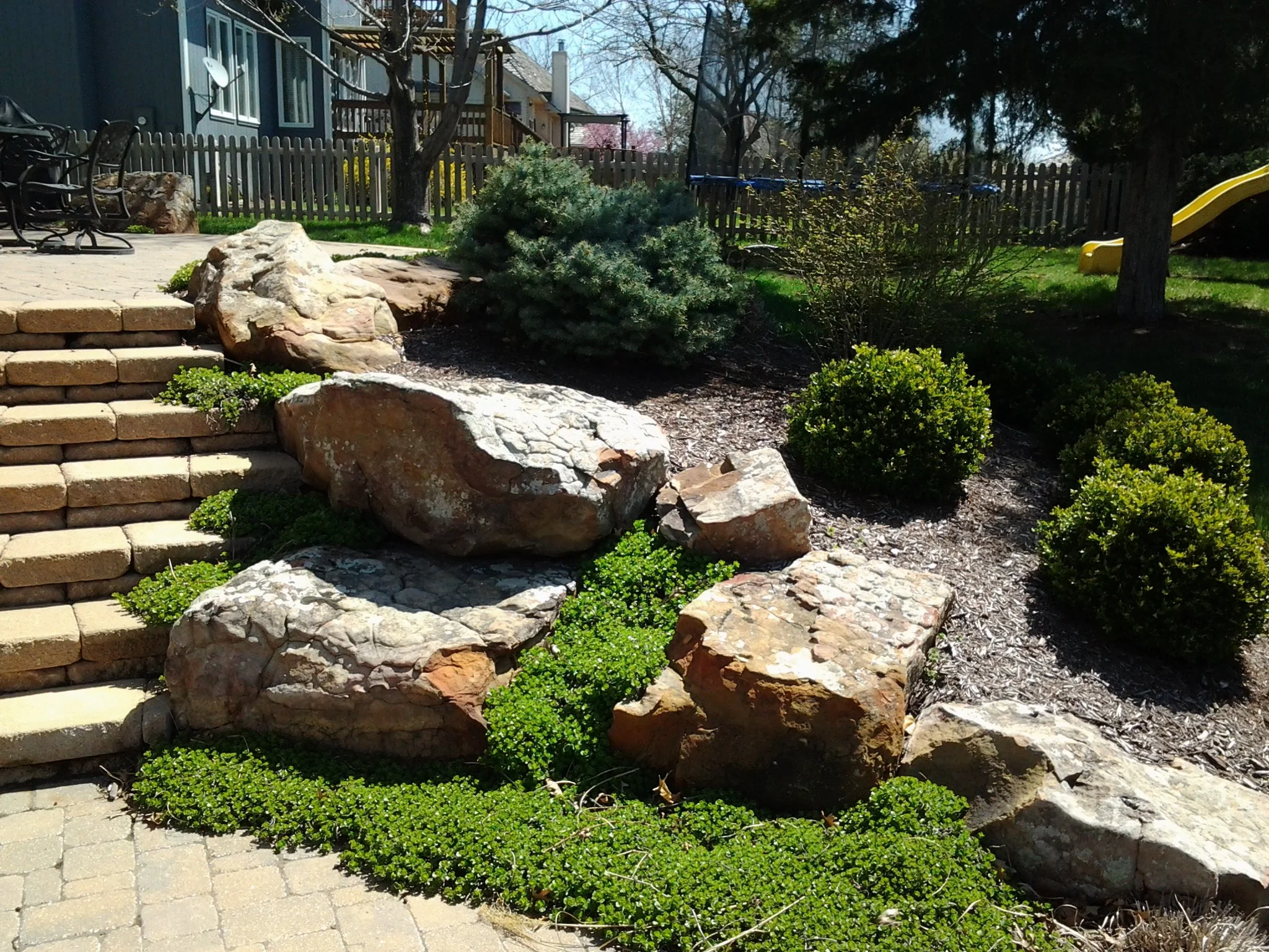 A landscaped backyard with stone steps, large rocks, green bushes, and a play area with a yellow slide in the background.