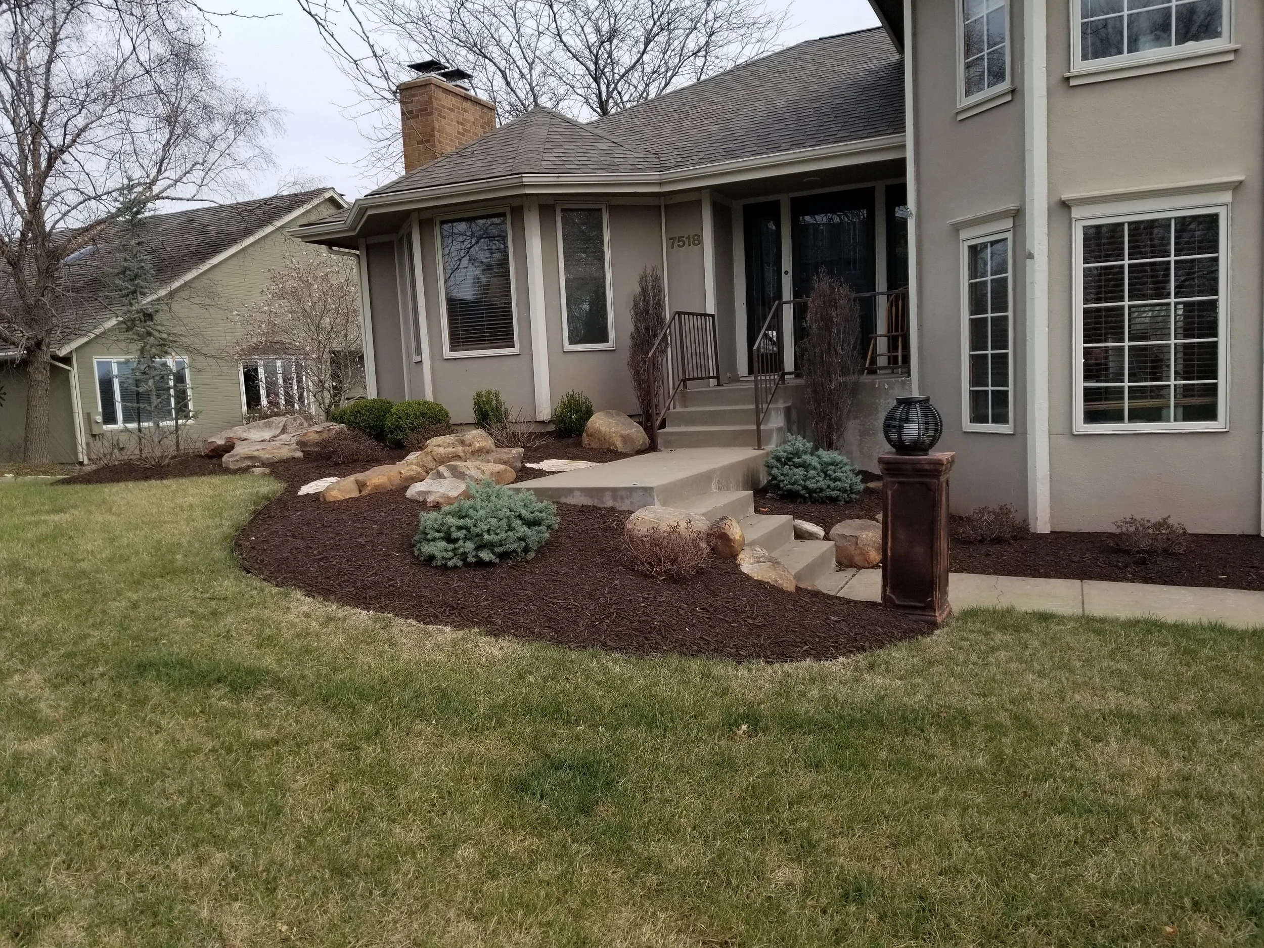 Front yard with green lawn, landscaped garden with rocks and shrubs, concrete steps leading to house entrance, house with beige exterior, multiple windows, and the address 7518 above the door, overcast sky.