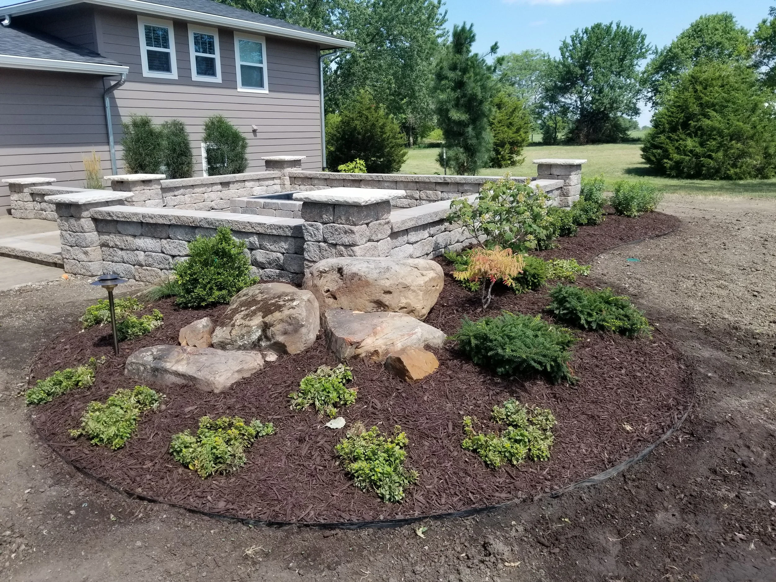 A landscaped garden bed featuring small shrubs, decorative rocks, and a stone retaining wall behind the plants. The garden is near a gray house with multiple windows.