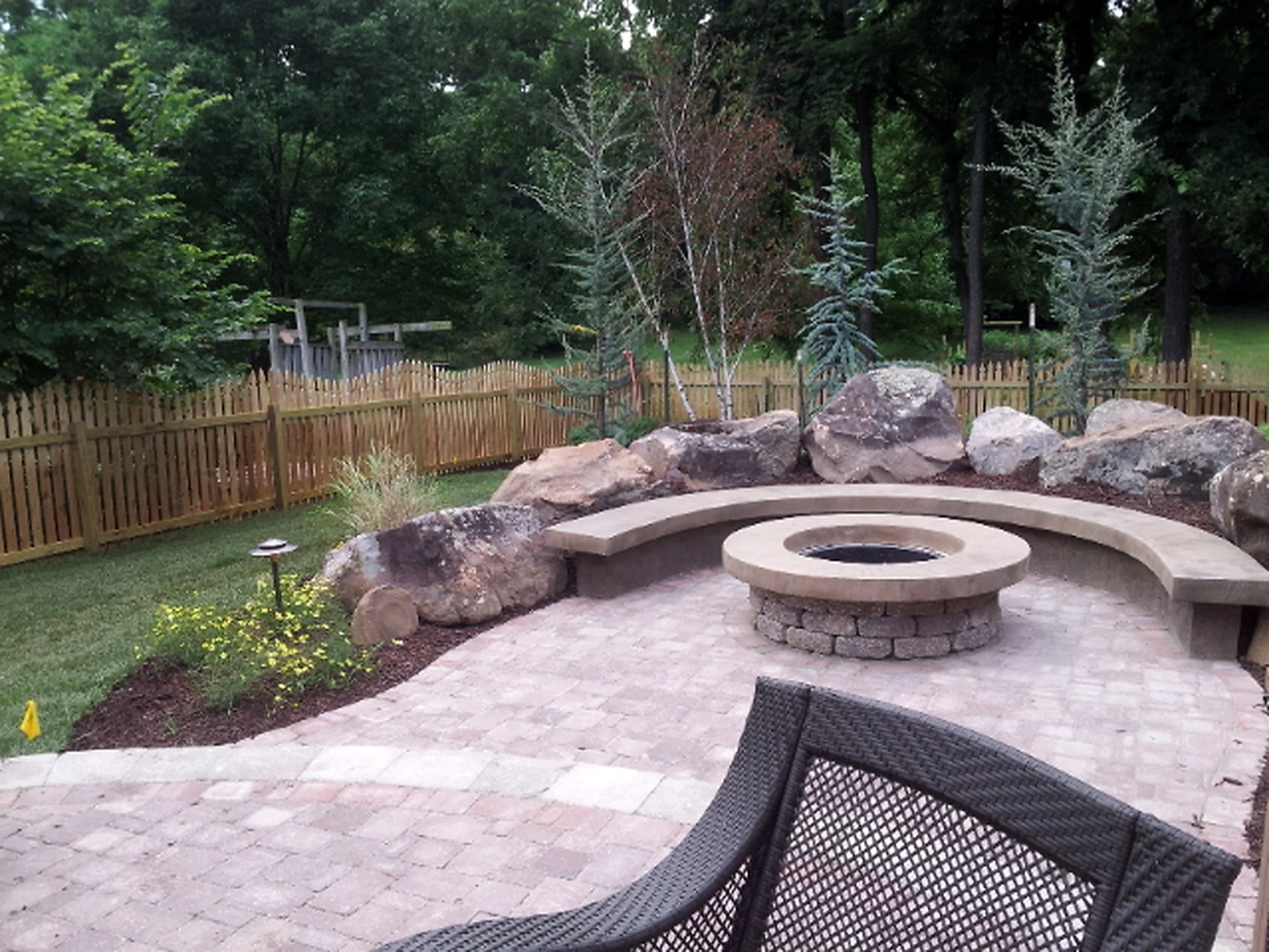 Backyard with a fire pit and curved bench, surrounded by rocks and trees, with a wooden fence in the background.
