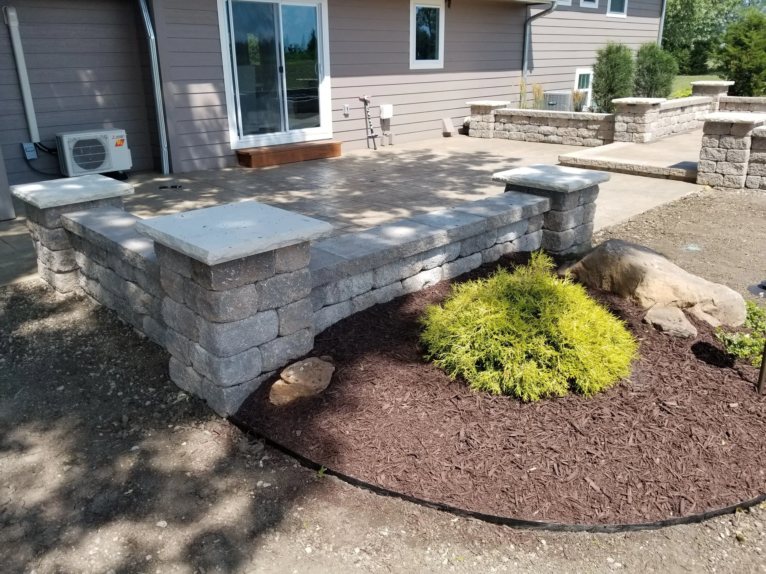 A backyard patio with a low stone wall and steps made of gray stone blocks, a sliding glass door, a small shrub, and large decorative rocks.