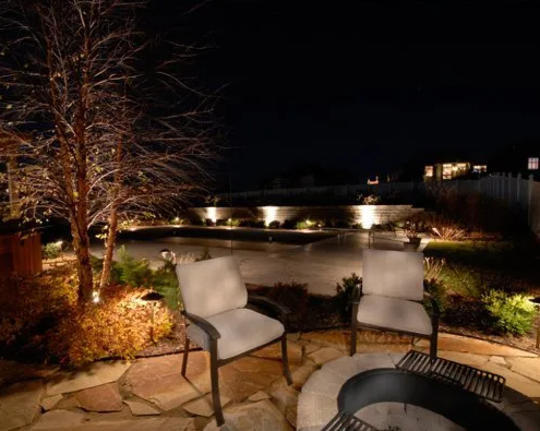 Backyard patio at night with two chairs, a fire pit, and decorative lighting along a pond or small pool.