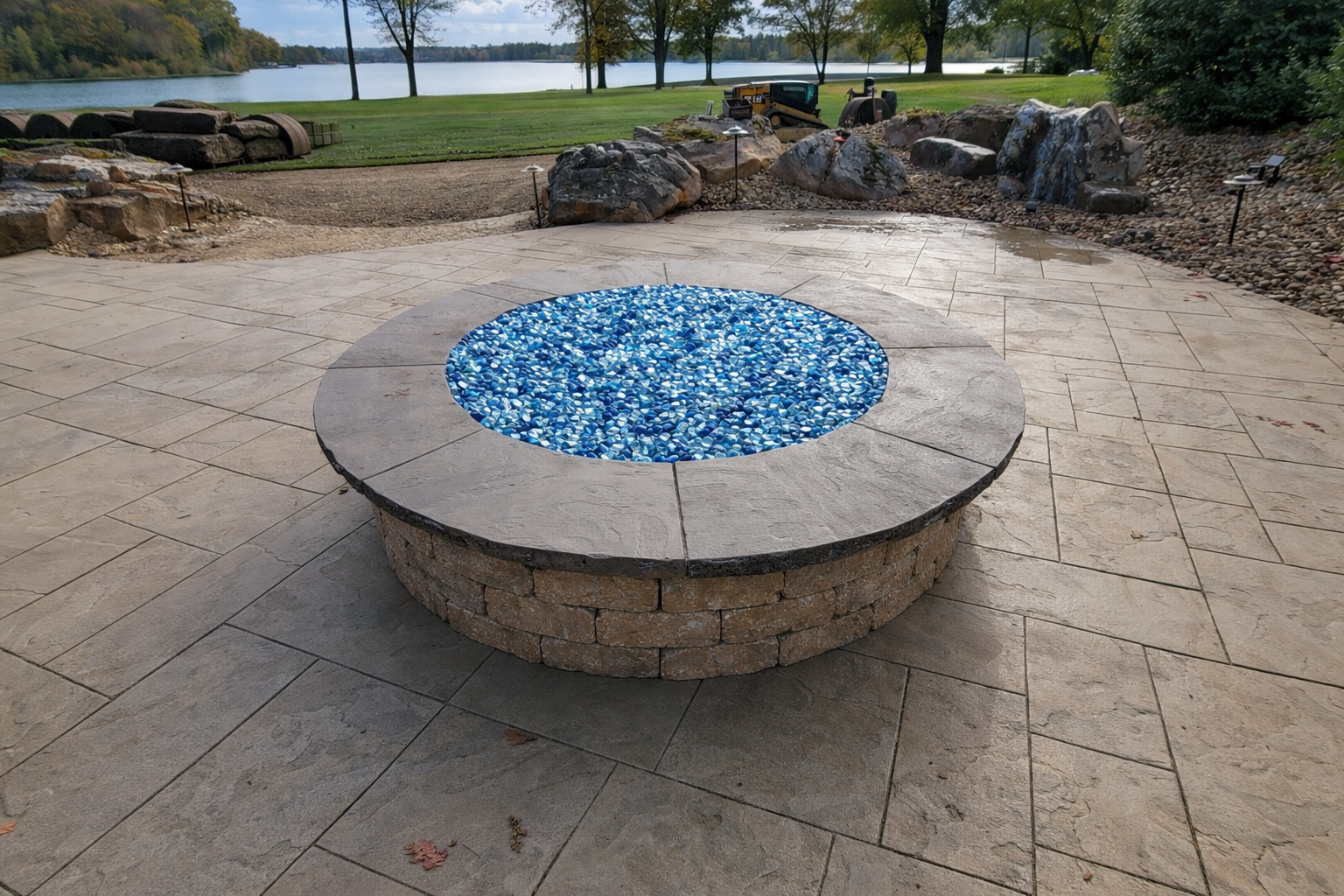 A circular fire pit with a stone outer wall and a top layer of large flat stones, filled with blue and white glass stones, set on a beige stone patio near a lake, with trees and rocks in the background.