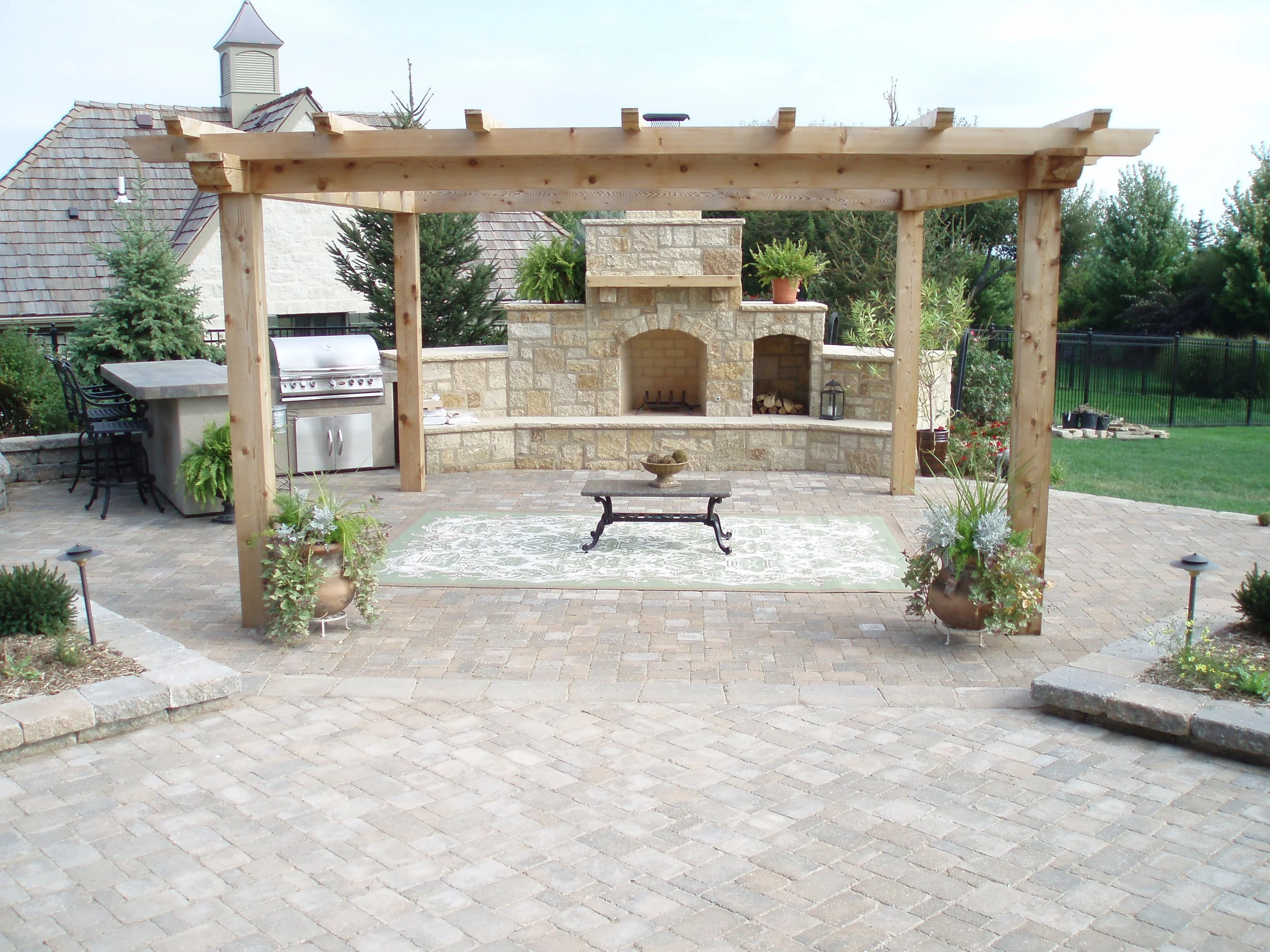 A backyard patio with a brick oven, a barbecue grill, a pergola frame, potted plants, a stone table, and outdoor seating, surrounded by a lawn and trees.