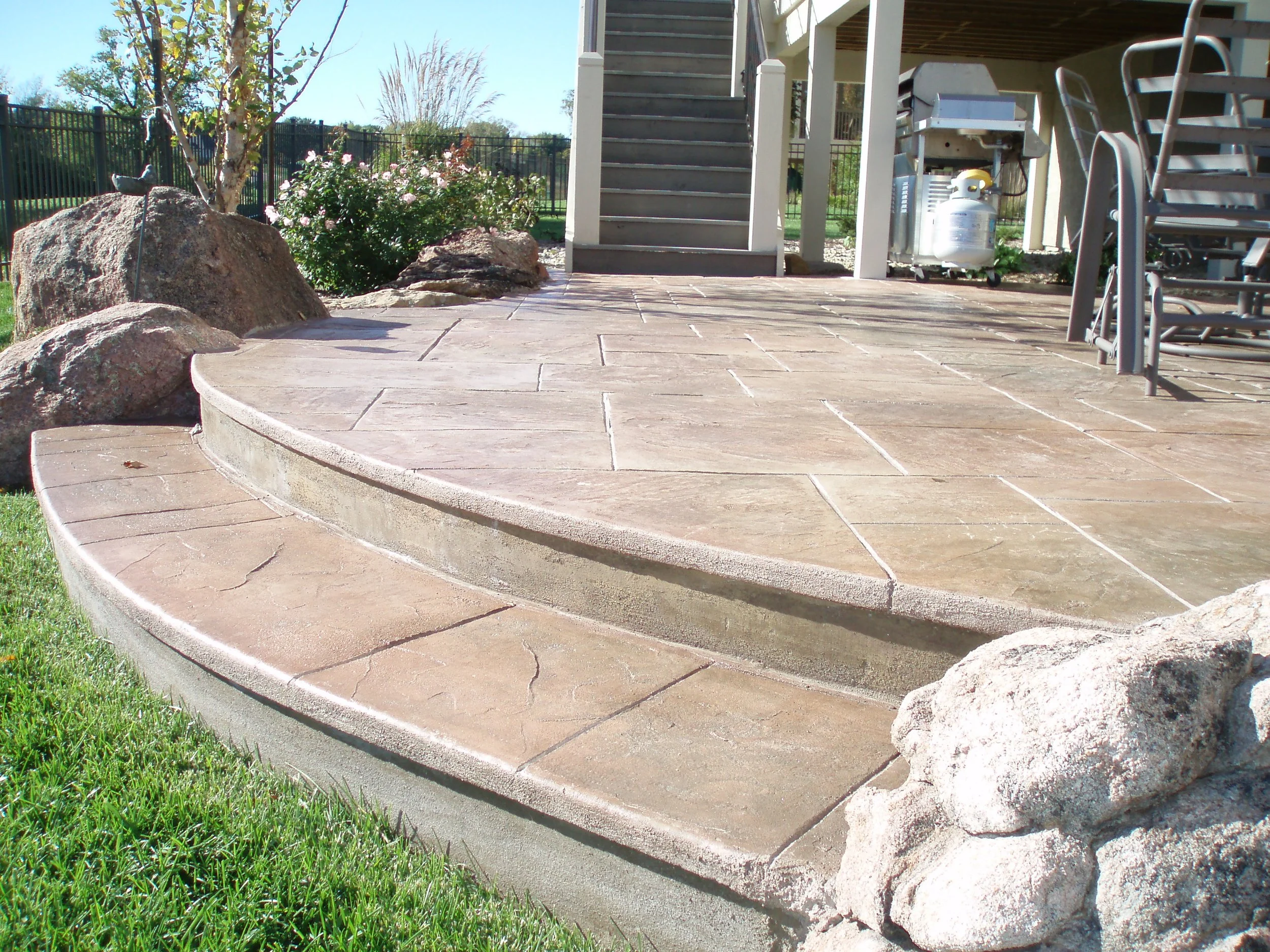 A patio with a curved concrete step, tan stone surface, surrounded by rocks and landscaping, with outdoor furniture, stairs, and grill in the background.
