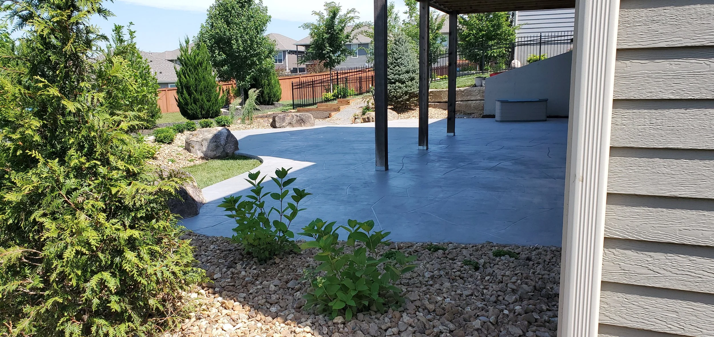 A backyard with a concrete patio, surrounded by rocks and plants, including shrubs and trees, with a wooden privacy fence and neighboring houses in the background.