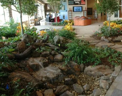Indoor garden with rocks, plants, and a flowing waterfall, adjacent to a seating area with tables, chairs, and a reception desk.