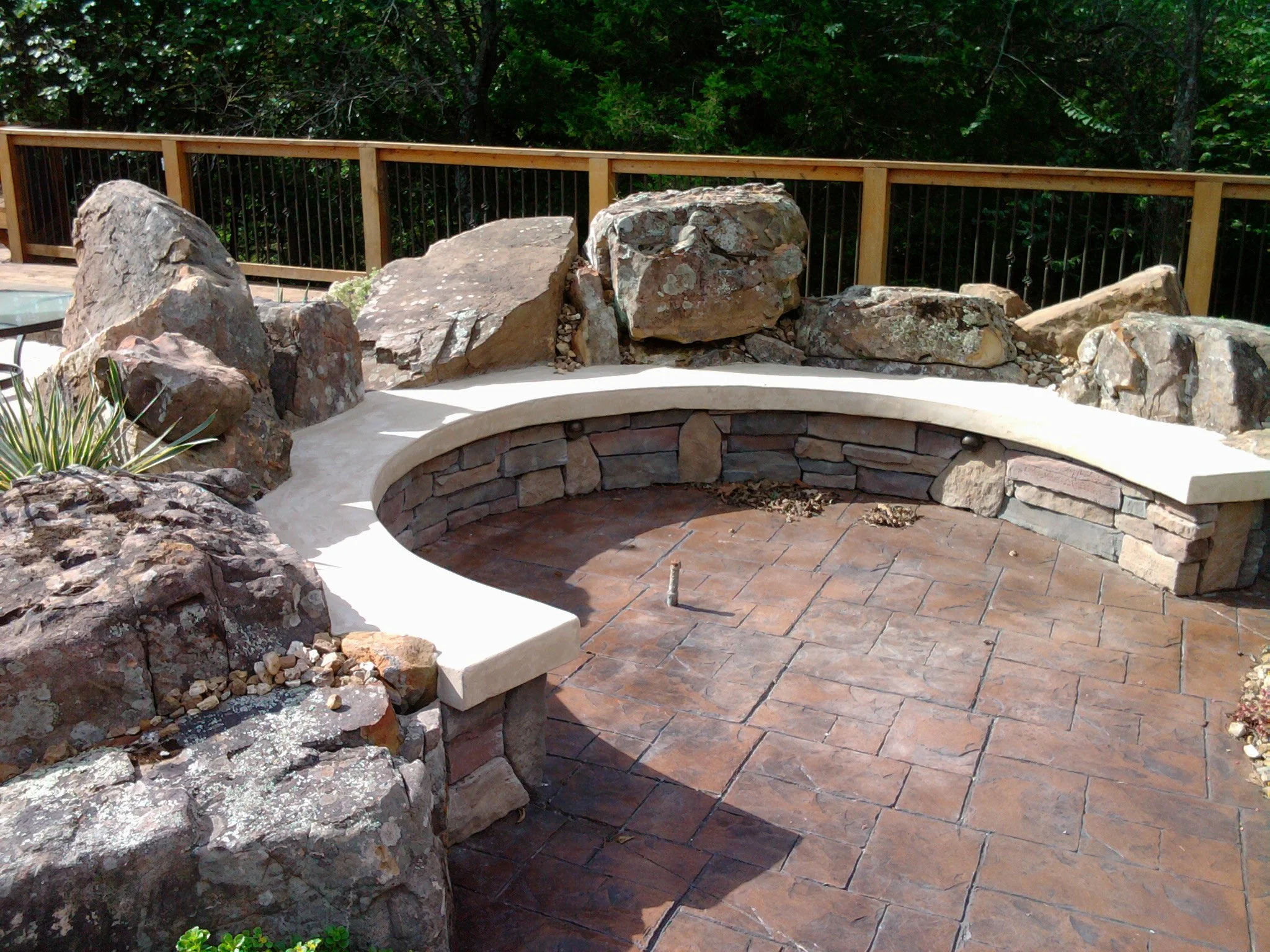 A curved stone fire pit with a brick interior, surrounded by large rocks, a concrete bench, and a wooden fence in a backyard with trees.
