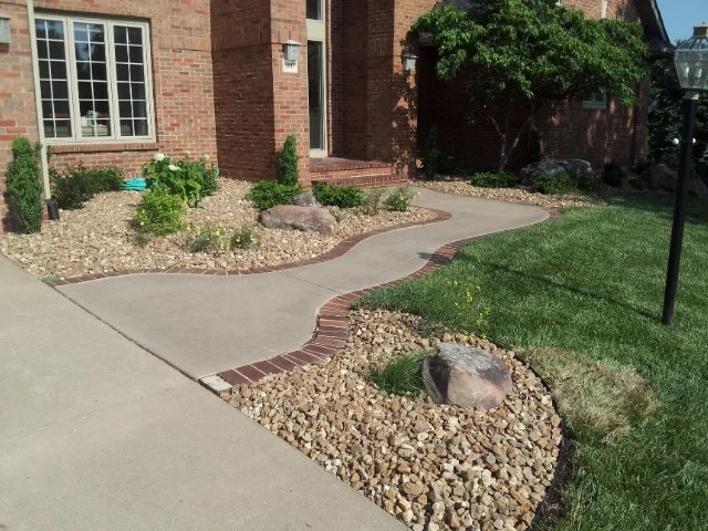 Curved concrete sidewalk with brick edging, surrounded by rock mulch and small plants in front of a brick house with a window and green lawn.