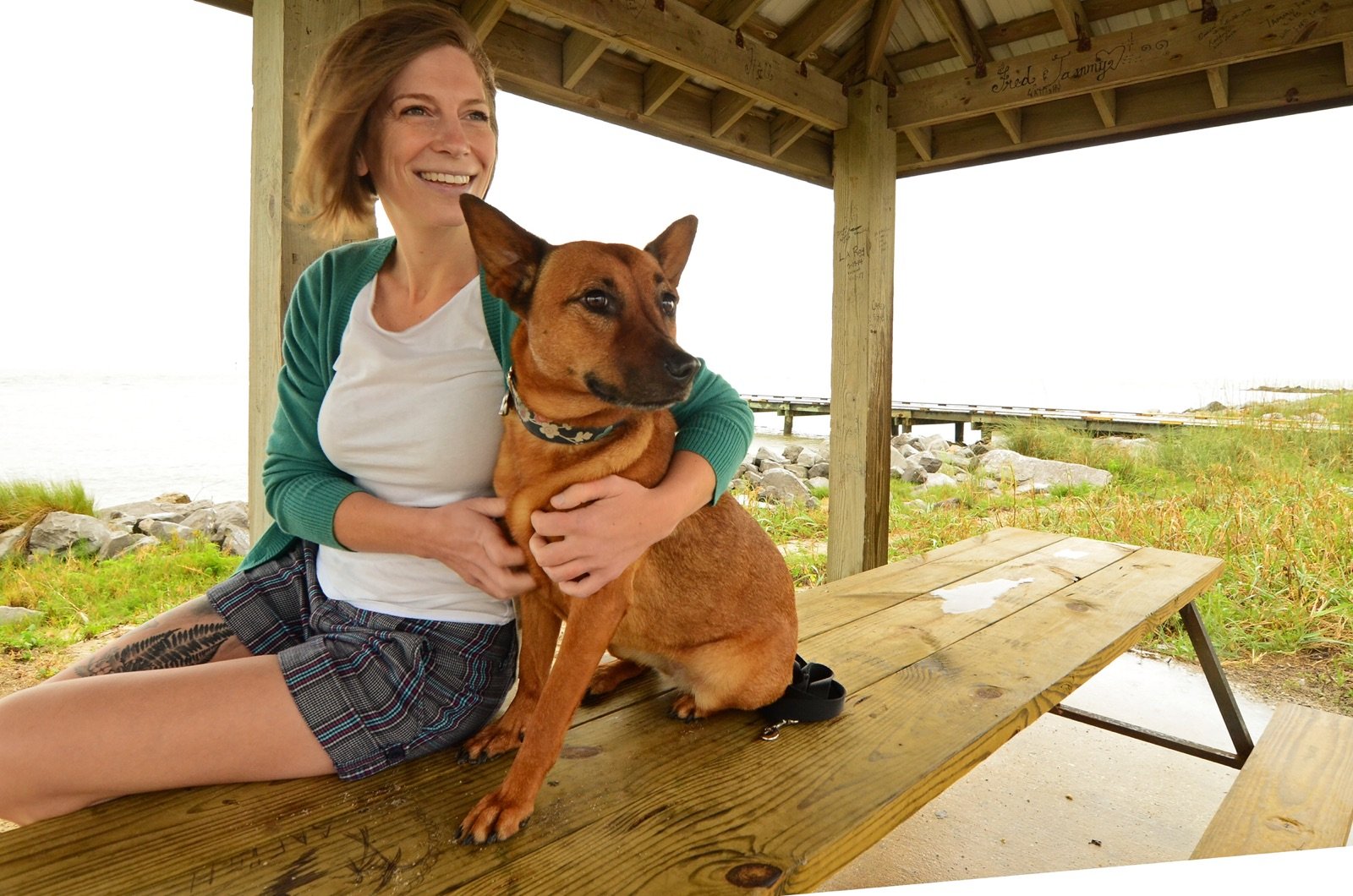 A woman smiling and sitting on a wooden picnic table, holding a brown dog under a wooden shelter near a body of water and greenery.