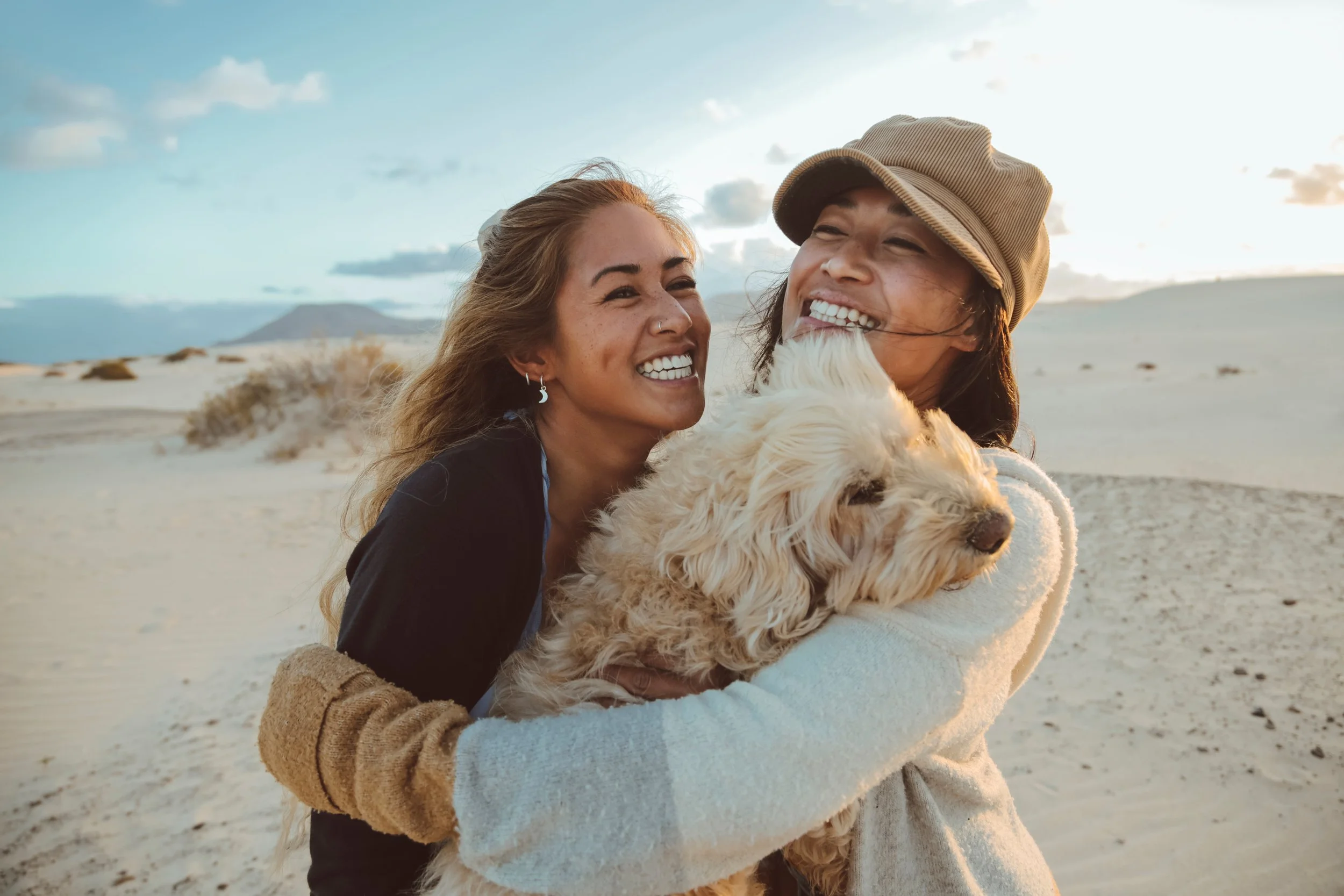 Two women smiling and hugging a fluffy goldendoodle dog on a sandy beach with mountains in the background, under a partly cloudy sky.
