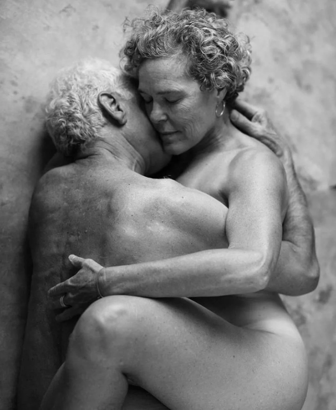 Naked older man and woman embracing each other with eyes closed, in a black and white photograph.