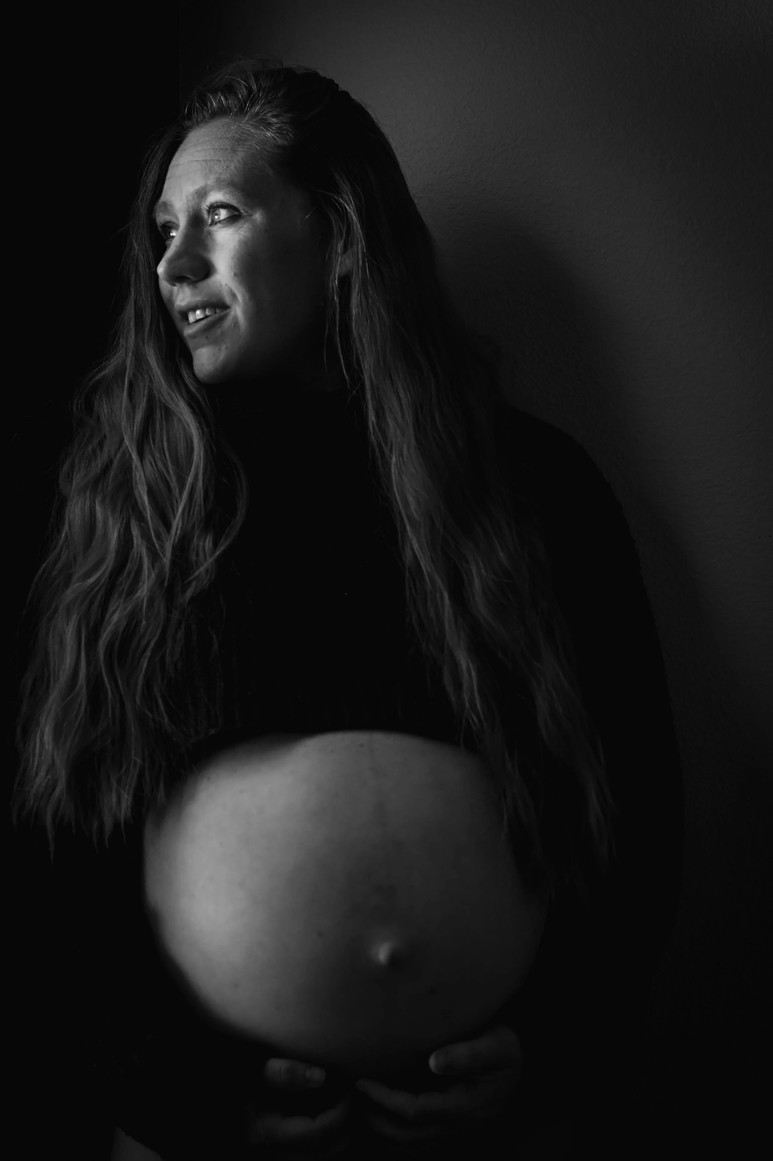 Black and white portrait of a pregnant woman with long wavy hair, looking to the side, holding her pregnant belly with both hands, against a plain wall.