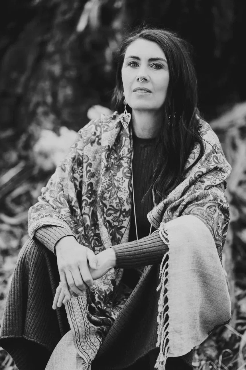 A woman with long dark hair, wearing a patterned shawl and knitted sweater, sits outdoors with trees in the background, looking directly at the camera in a black and white photograph.