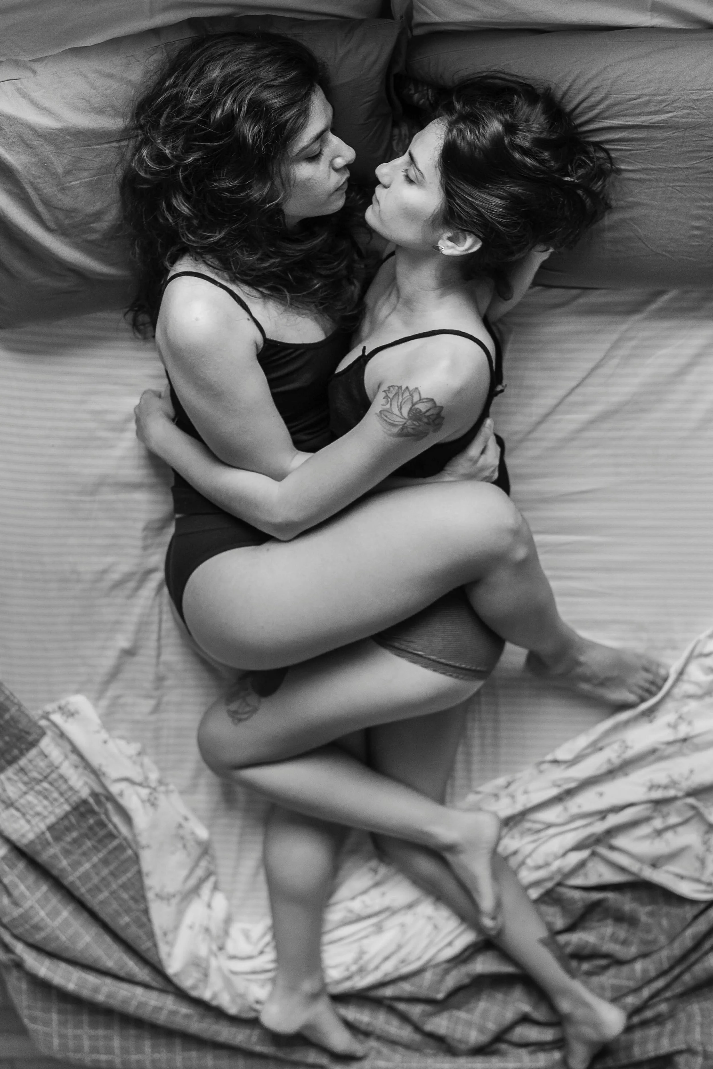 Two women lying on a bed, facing each other, embracing, in black and white. They are wearing black lingerie, and one has a tattoo on her upper arm.