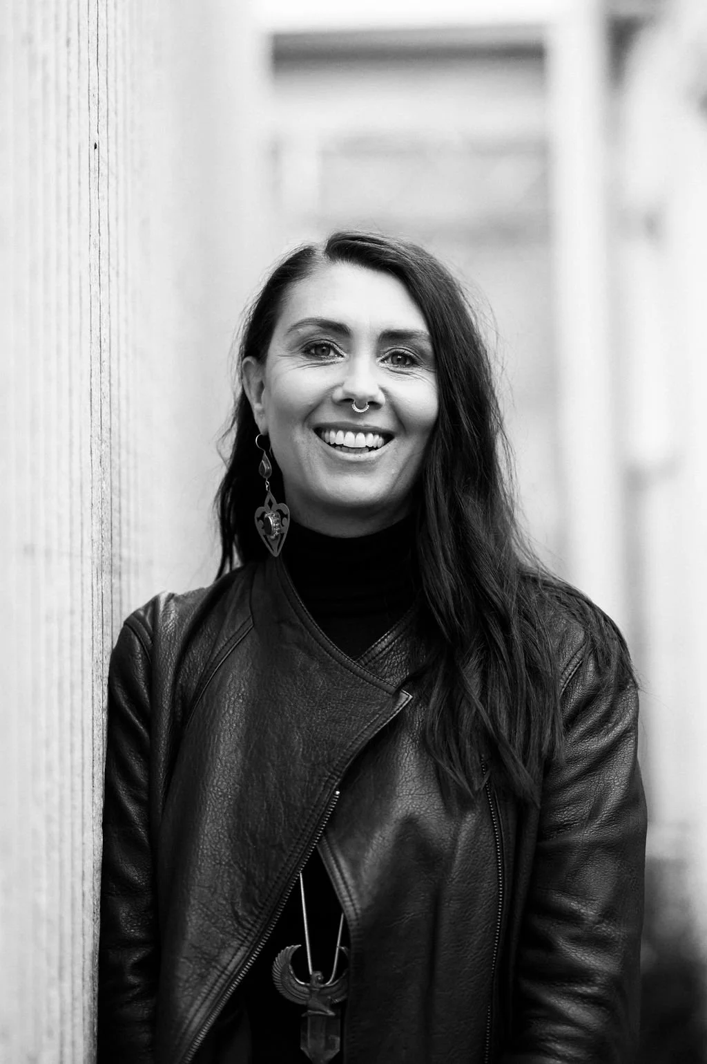 A woman with dark hair and a septum piercing, smiling, wearing earrings, a black top, and a leather jacket, standing against a wall in an outdoor setting.