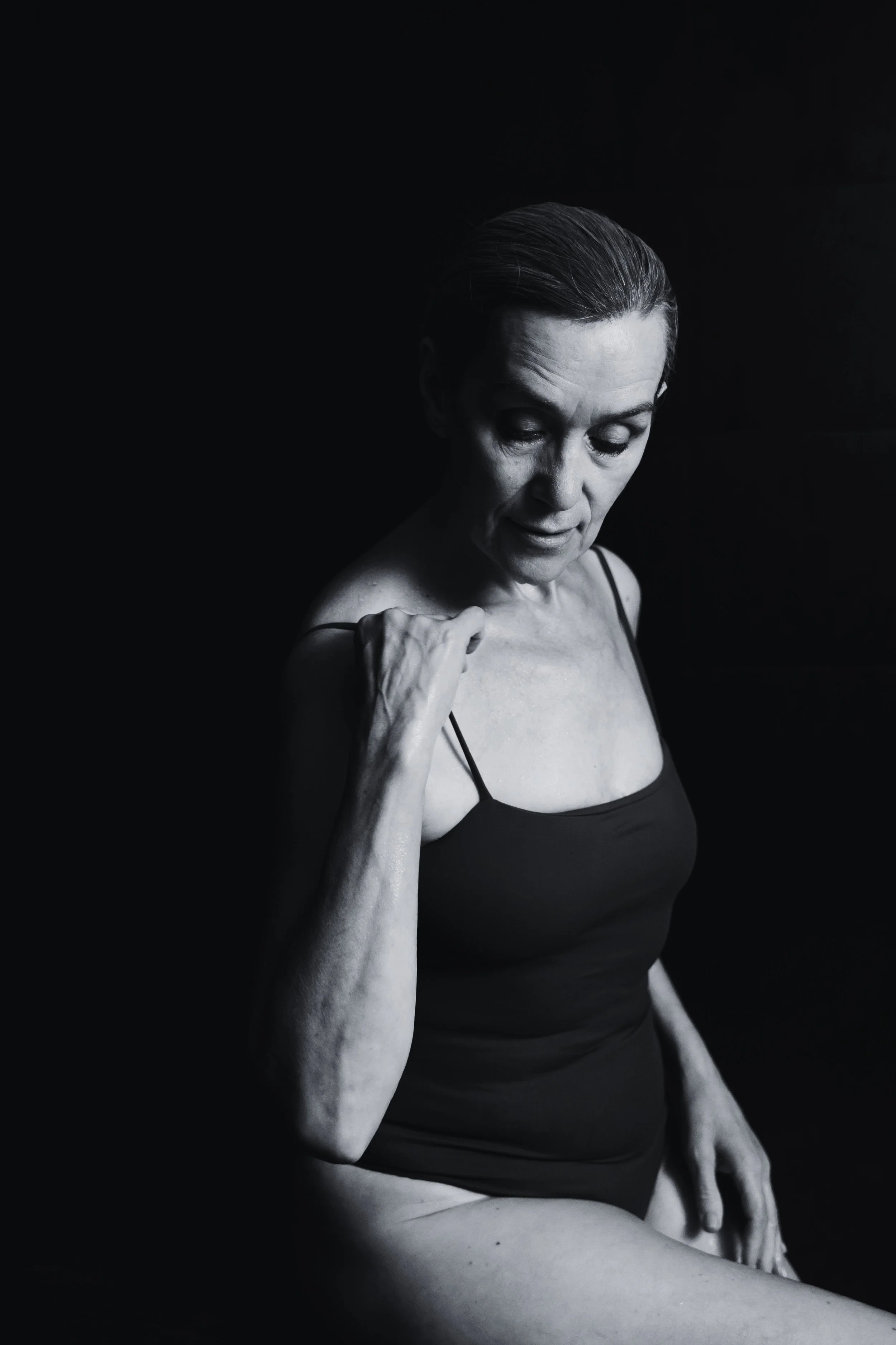 A black and white photo of an elderly woman in a black tank top, sitting with her head bowed, one hand holding her shoulder, with a dark background.
