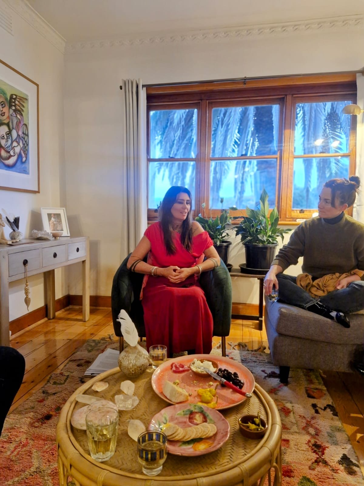 Two women sitting in a living room, having a conversation. A round rattan table in front holds food and drinks, with a window showing a view of trees in the background.