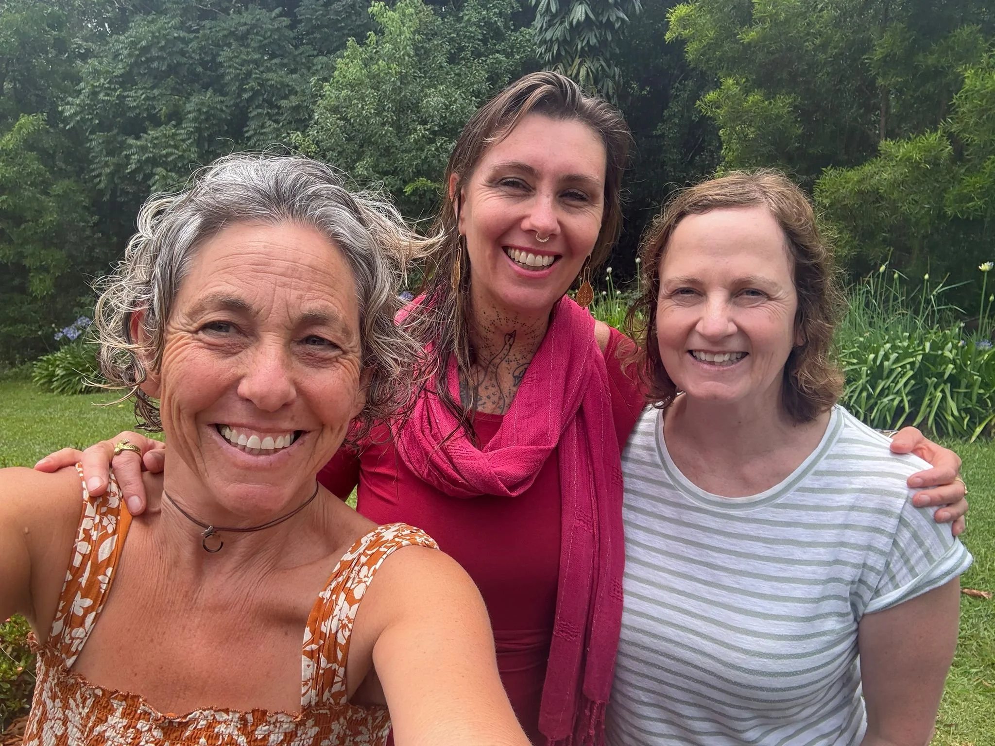 Three women smiling and taking a selfie outdoors in a lush garden.