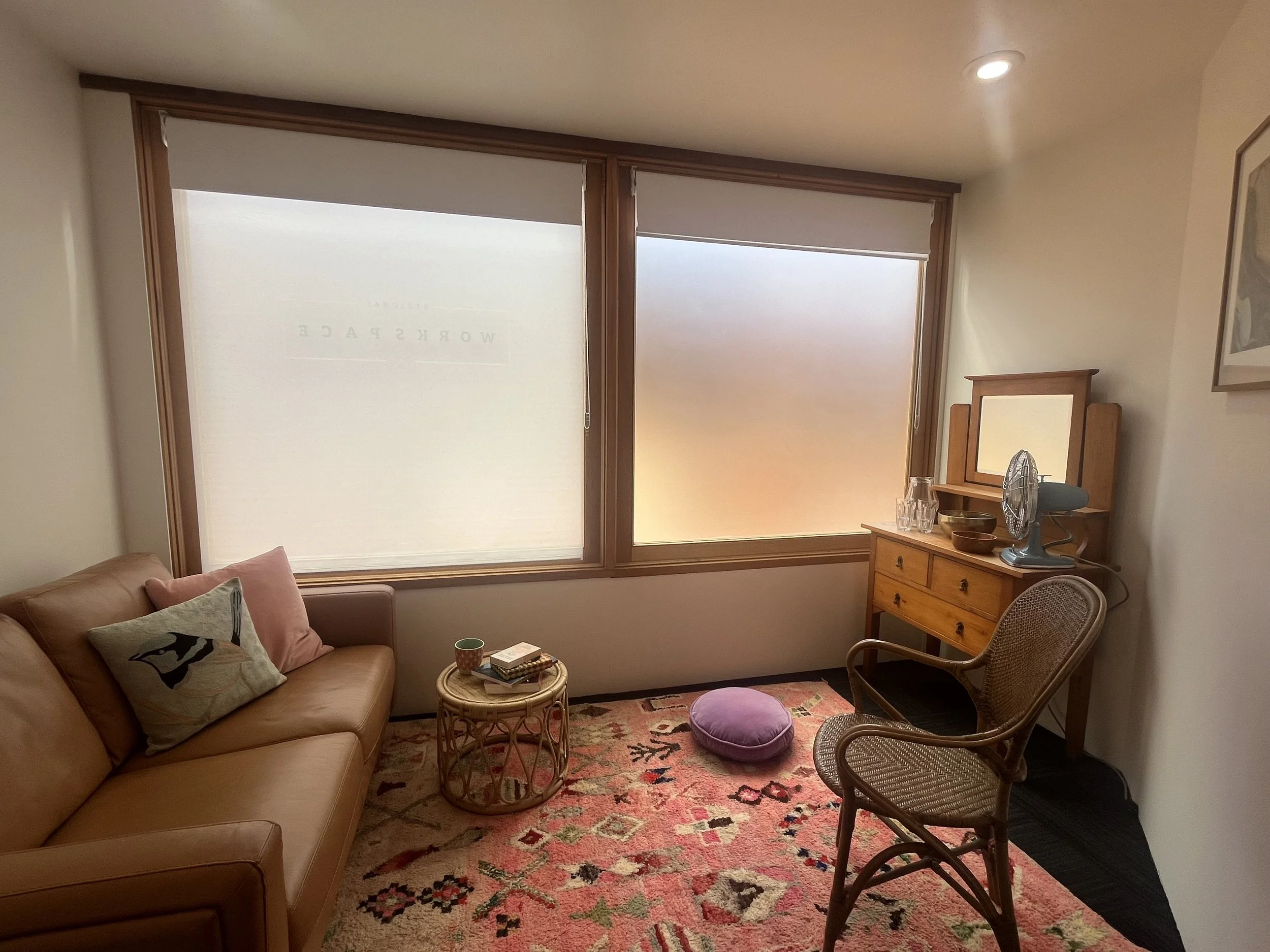 A cozy living room with large windows covered by white blinds. It features a tan leather sofa with pillows, a pink and a patterned cushion. In front of the sofa is a small wicker table with books, a cup, and plates. To the side, there's a wooden dresser with a mirror and a teal fan, along with glassware. A rattan chair is placed nearby, and a pink patterned rug covers the floor.